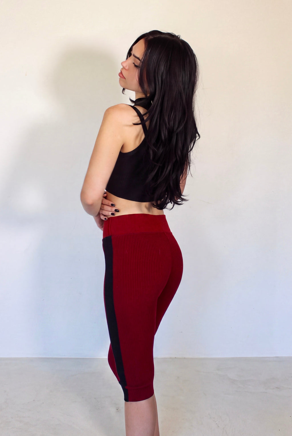 A woman stands in profile against a plain off-white wall, wearing a black sports bra and dark red high-waisted capri leggings with black side panels.