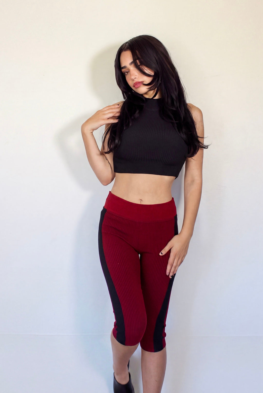 A woman modeling a two-piece set consisting of a black mock-neck crop top and dark red capri leggings with black side panels, standing against a plain off-white wall.