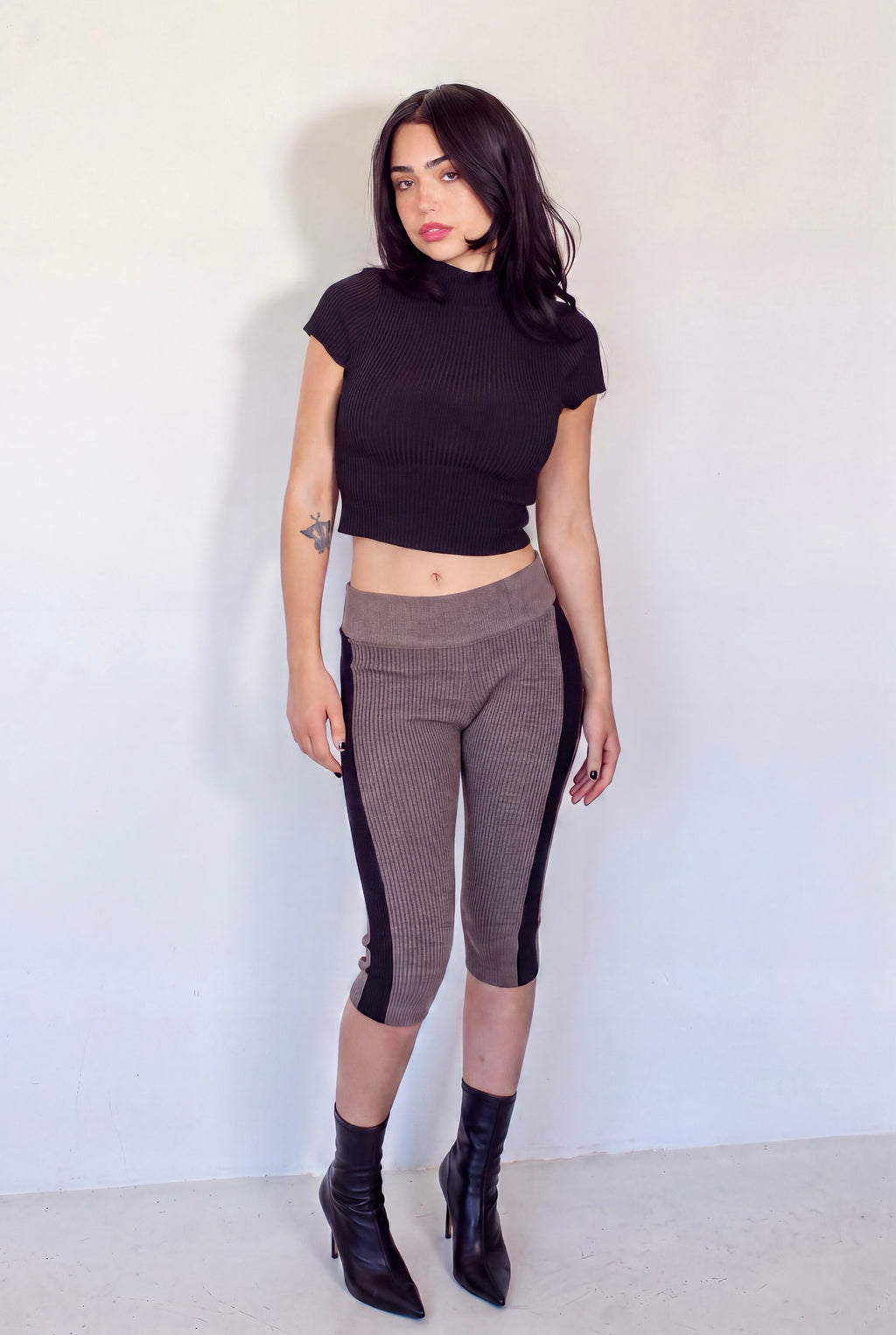 A woman stands against a plain off-white wall wearing a black short-sleeve ribbed mock-neck crop top, brown ribbed capri leggings with black side panels, and black ankle boots.