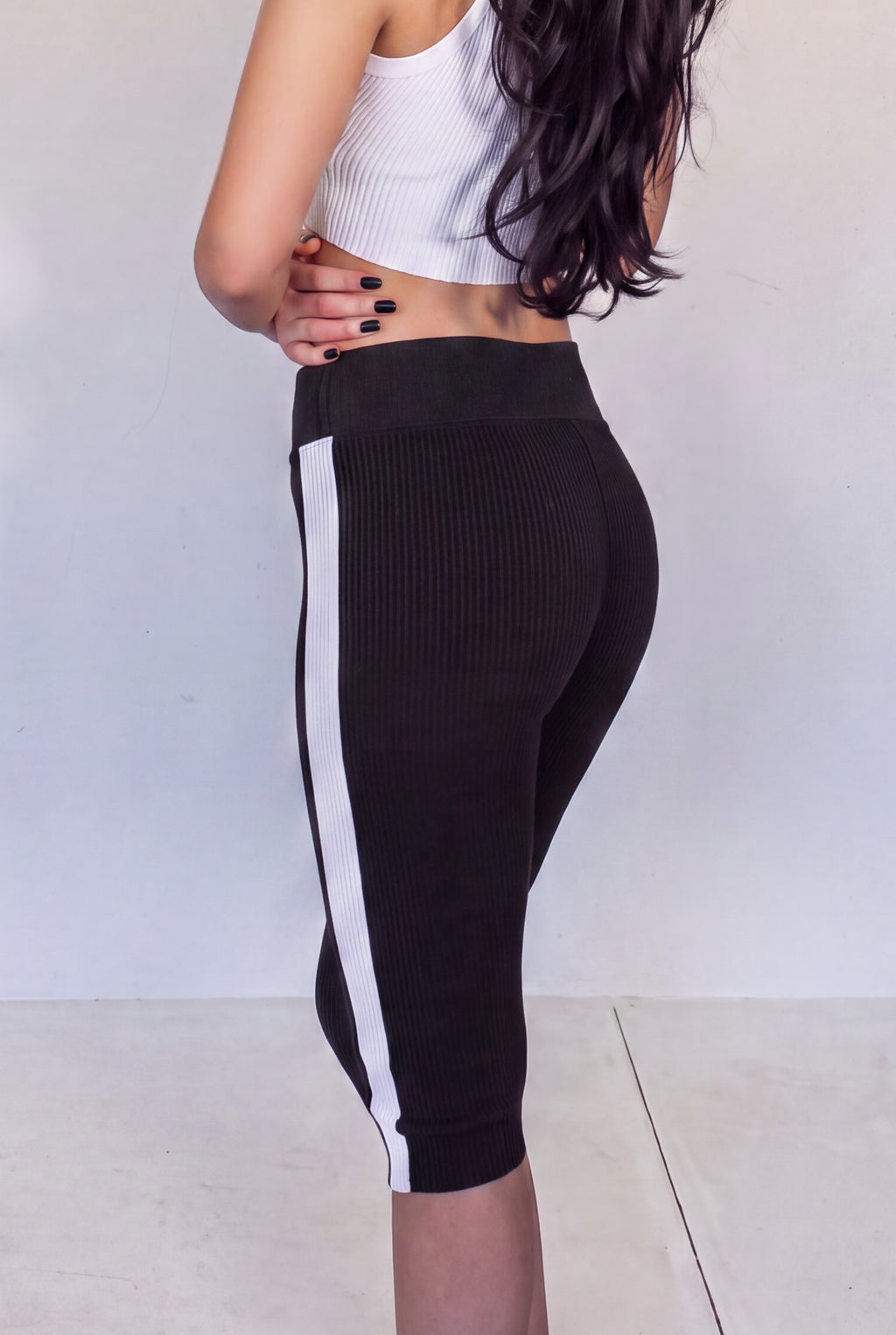 A woman wearing black knit ribbed capri leggings with a bold white side stripe, shown from a side-back angle against a plain light gray wall.