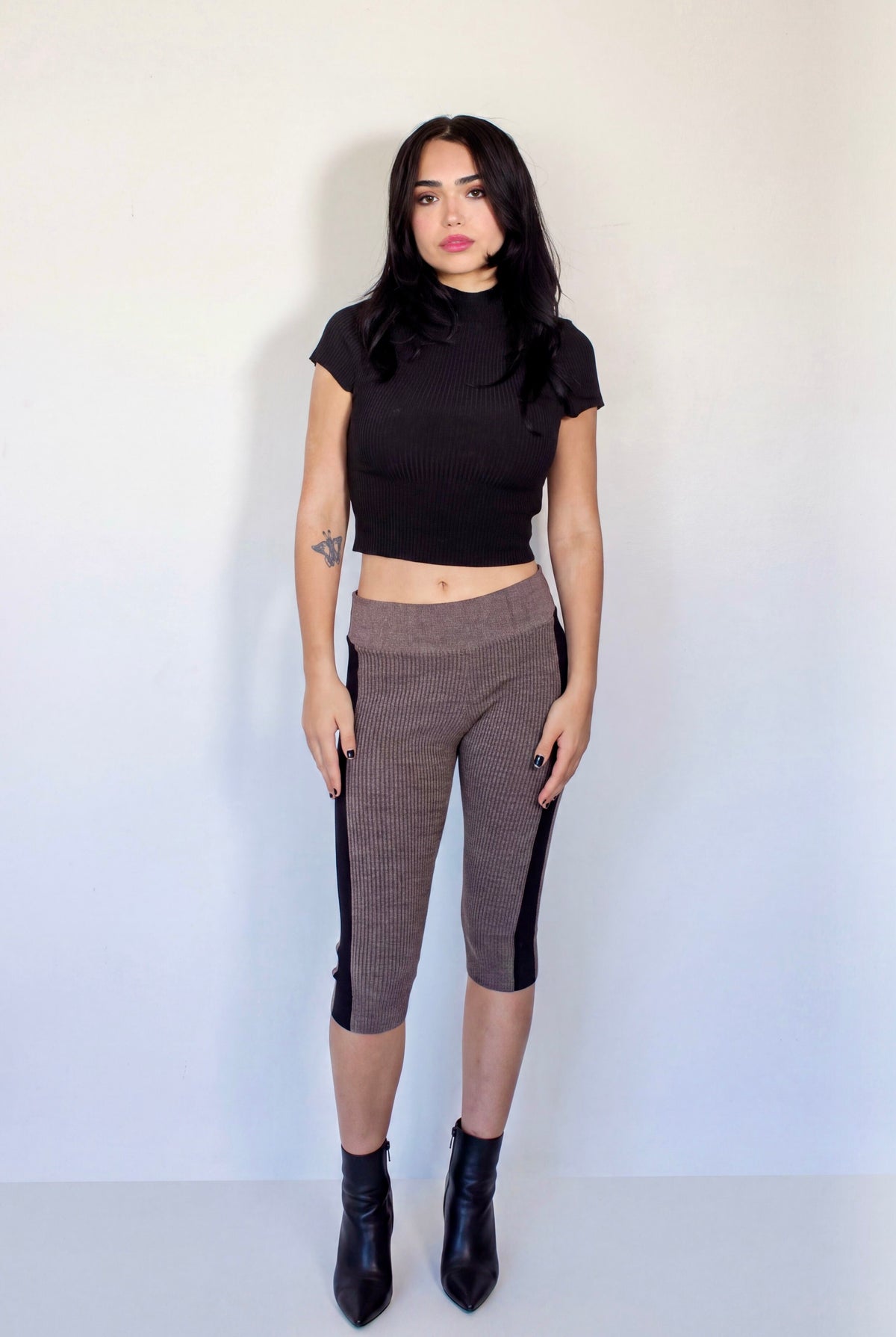 A woman stands against a plain off-white wall wearing a black short-sleeve crop top, brown ribbed capri pants with black side panels, and black ankle boots.