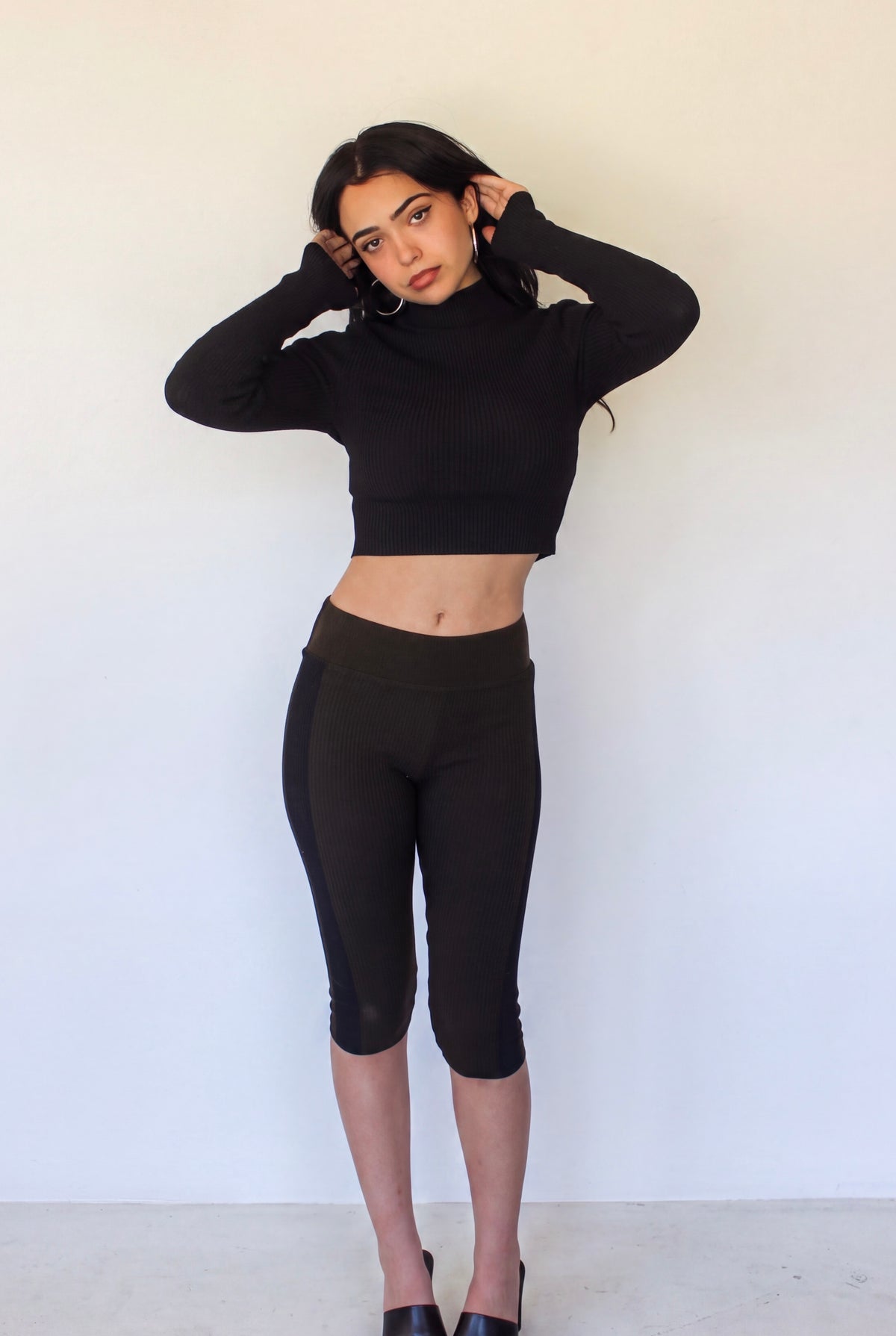 A woman modeling a black long-sleeve cropped turtleneck top and matching black capri leggings against a plain off-white wall.