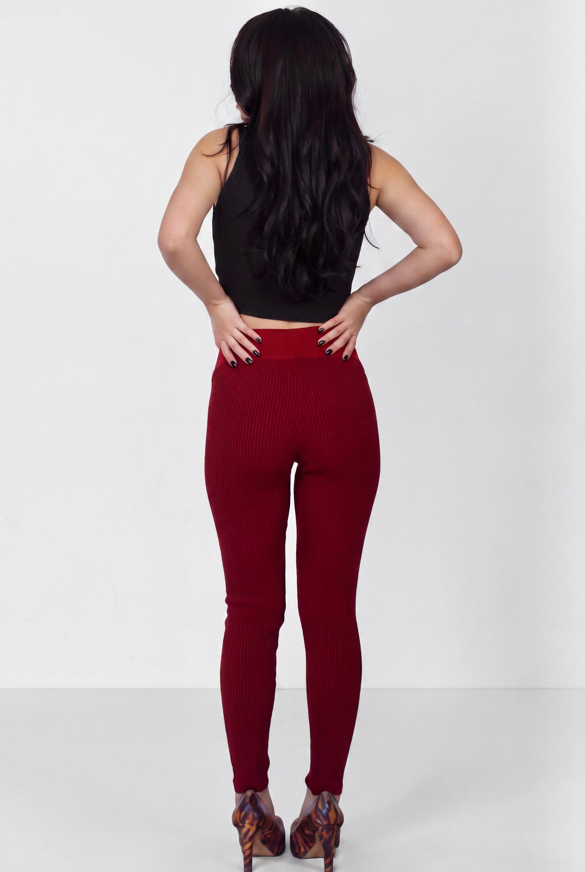 A woman shown from the back wearing a black sleeveless crop top and dark red ribbed leggings, standing against a plain light gray studio background.
