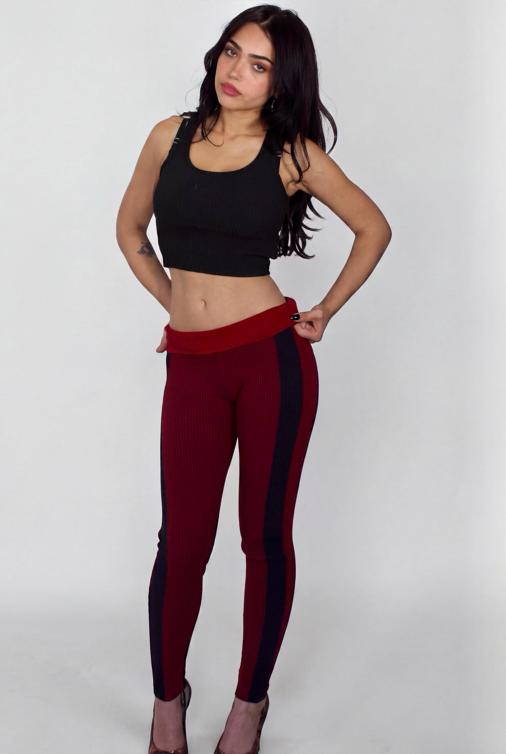 A woman modeling a pair of burgundy leggings with black side panels and a wide red waistband, paired with a black crop top, against a light gray studio background.