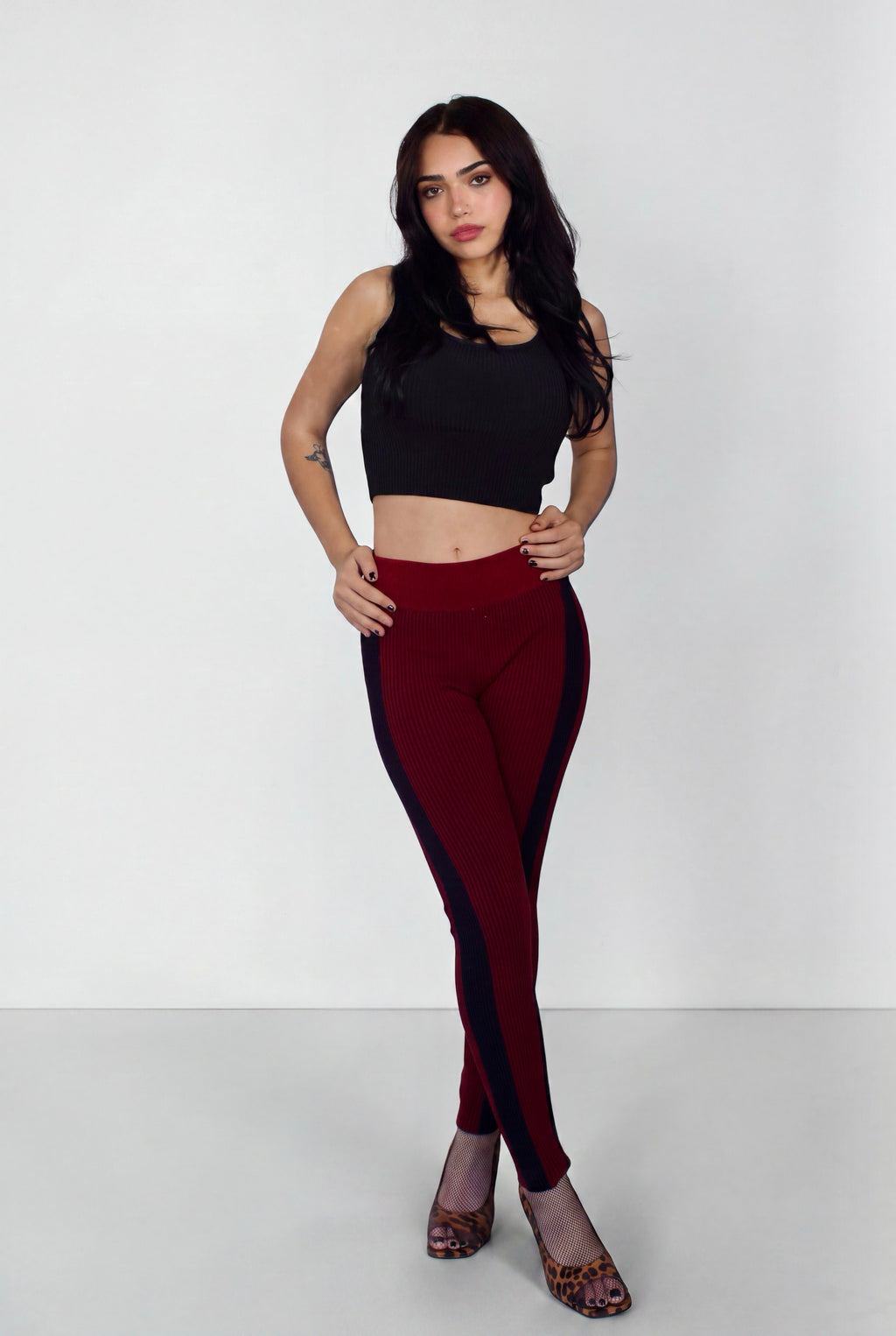 A woman modeling a pair of burgundy leggings with dark navy side panels, paired with a black crop top and leopard print heels, against a plain light gray studio background.