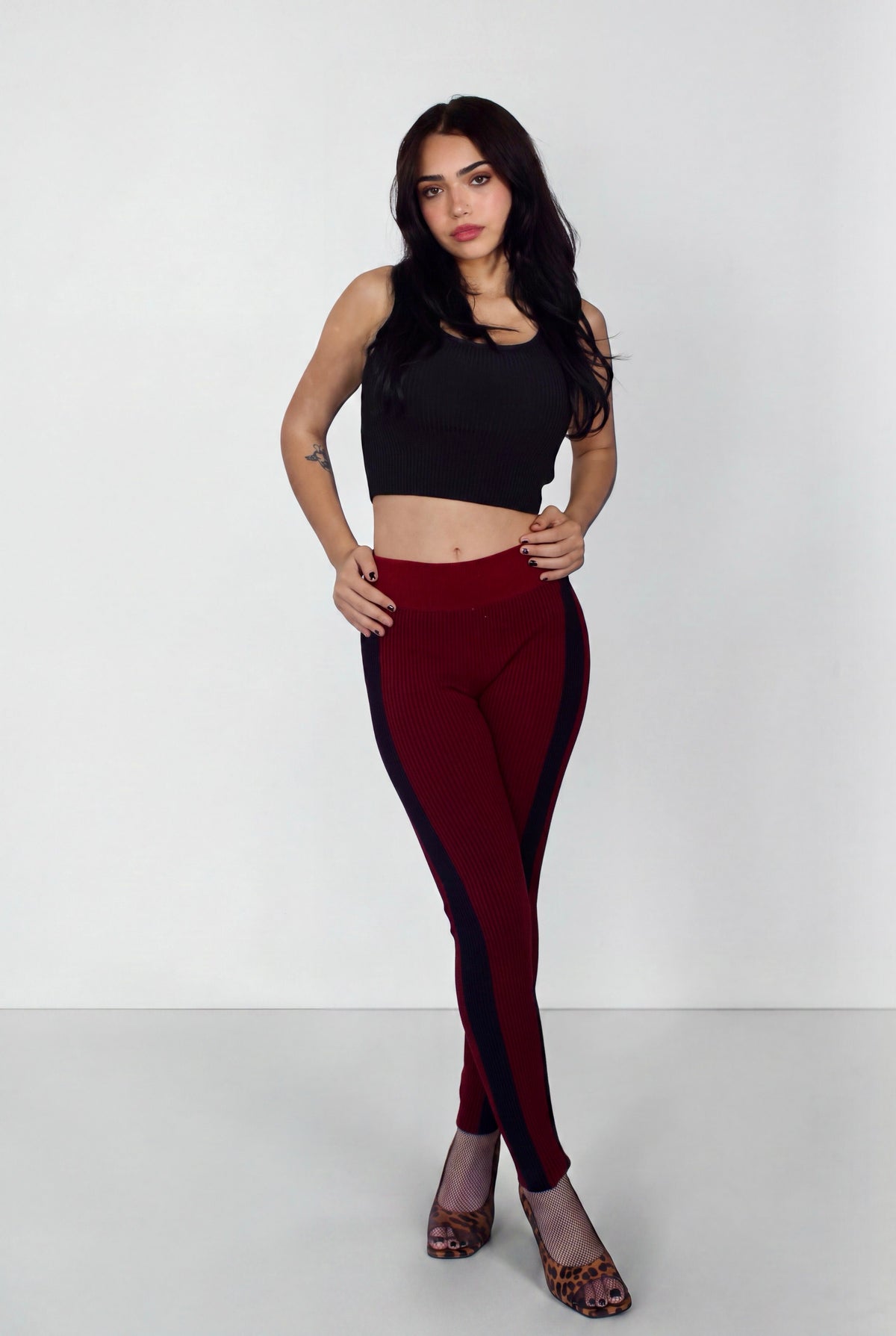 A woman modeling a pair of burgundy leggings with dark navy side panels, paired with a black crop top and leopard print heels, against a plain light gray studio background.