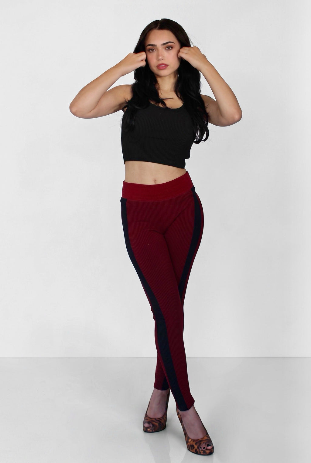 A woman modeling a pair of high-waisted leggings in deep burgundy with dark navy side panels, paired with a black crop top and patterned heels, against a plain white studio background.