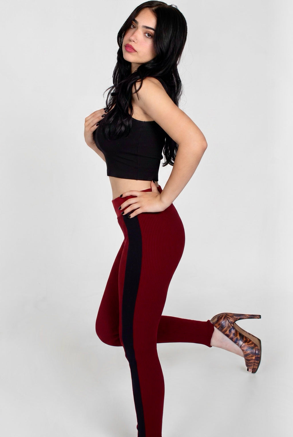 A woman posing in a black crop top and high-waisted burgundy leggings with a black side stripe, paired with patterned high heels, against a light gray studio background.