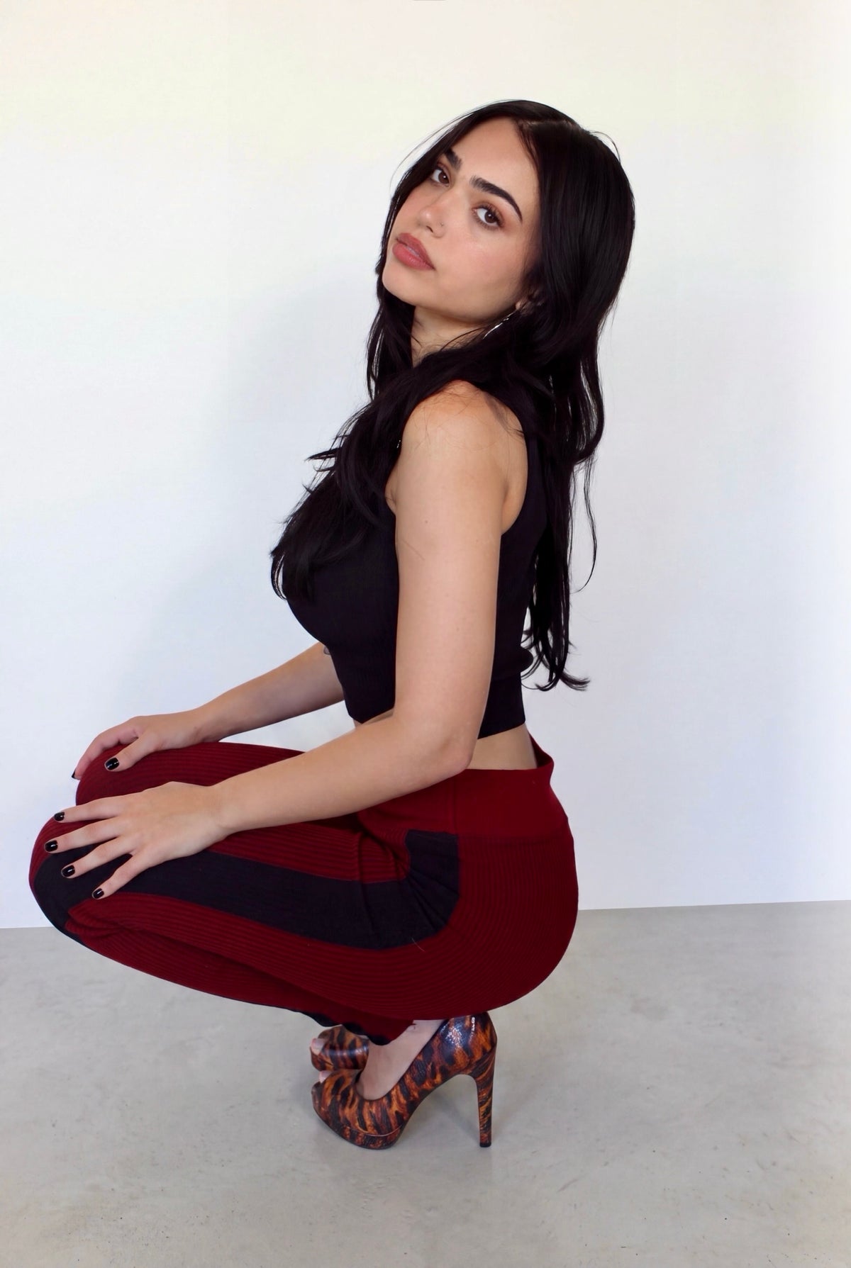 A woman crouching in a studio setting, wearing a black sleeveless crop top, deep red high-waisted pants with black side panels, and leopard-print high heels against a plain white wall and light gray floor.