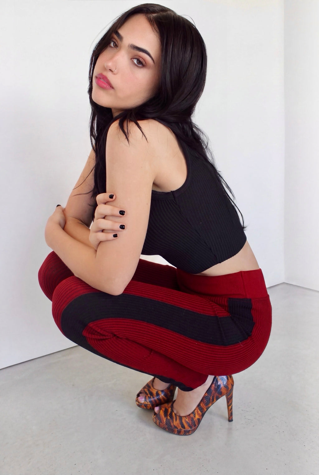 A woman crouching in a studio setting, wearing a black ribbed tank top and red-and-black striped ribbed leggings, paired with patterned high heels.