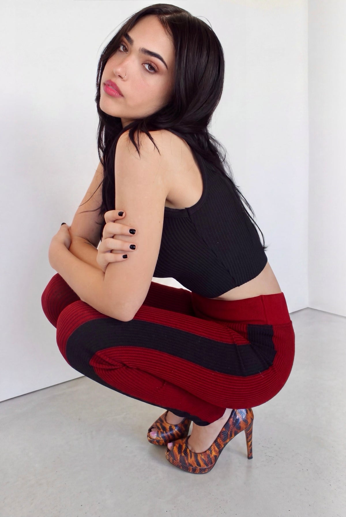 A woman crouching in a studio setting, wearing a black ribbed tank top and red-and-black striped ribbed leggings, paired with patterned high heels.