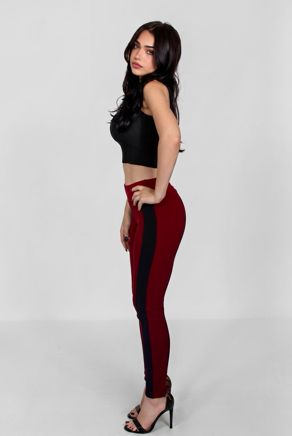 A woman modeling high-waisted, form-fitting leggings in deep burgundy with a black side panel, paired with a black crop top and black high-heeled sandals, set against a plain light gray studio background.