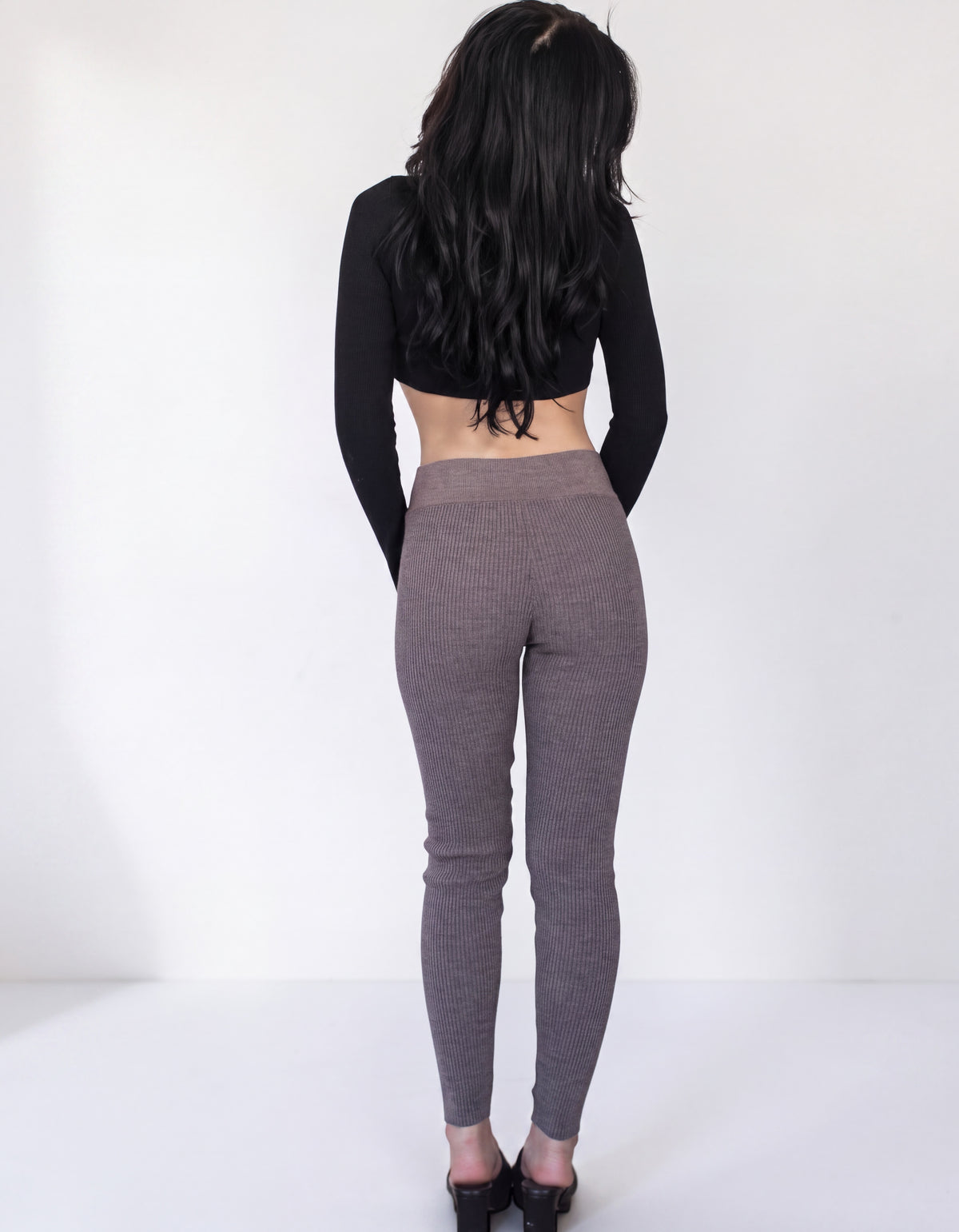 A woman viewed from behind wearing a black long-sleeve crop top and textured gray ribbed leggings, standing against a plain white studio background.