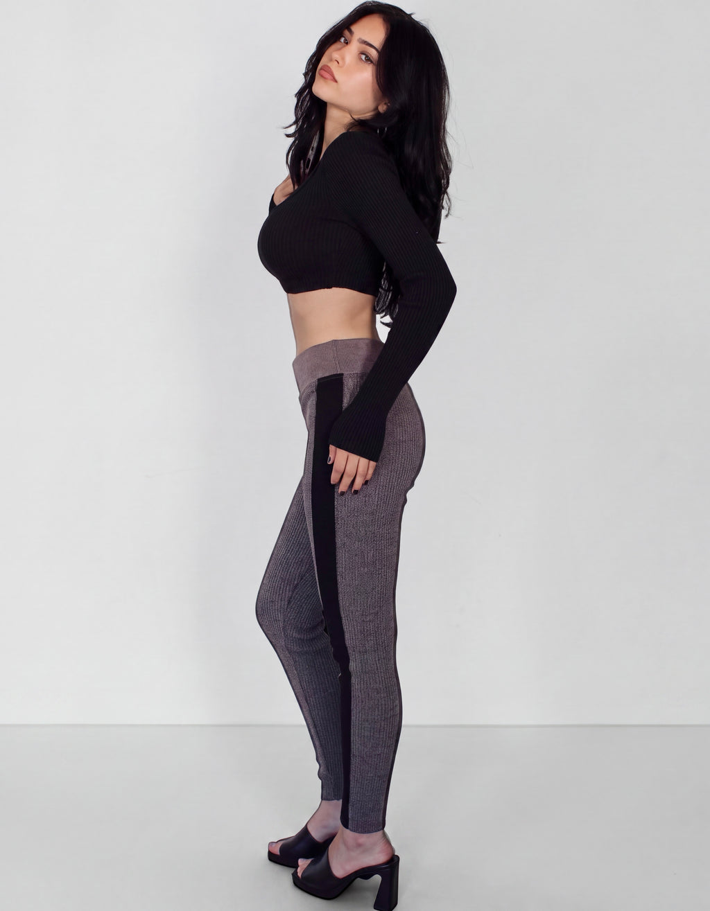A woman modeling a black long-sleeve crop top and gray ribbed leggings with black side panels, standing in a studio against a light gray background.