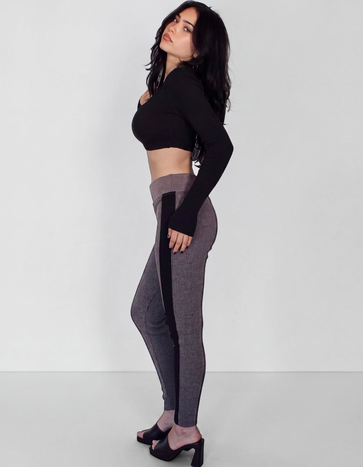 A woman modeling a black long-sleeve crop top and gray ribbed leggings with black side panels, standing in a studio against a light gray background.