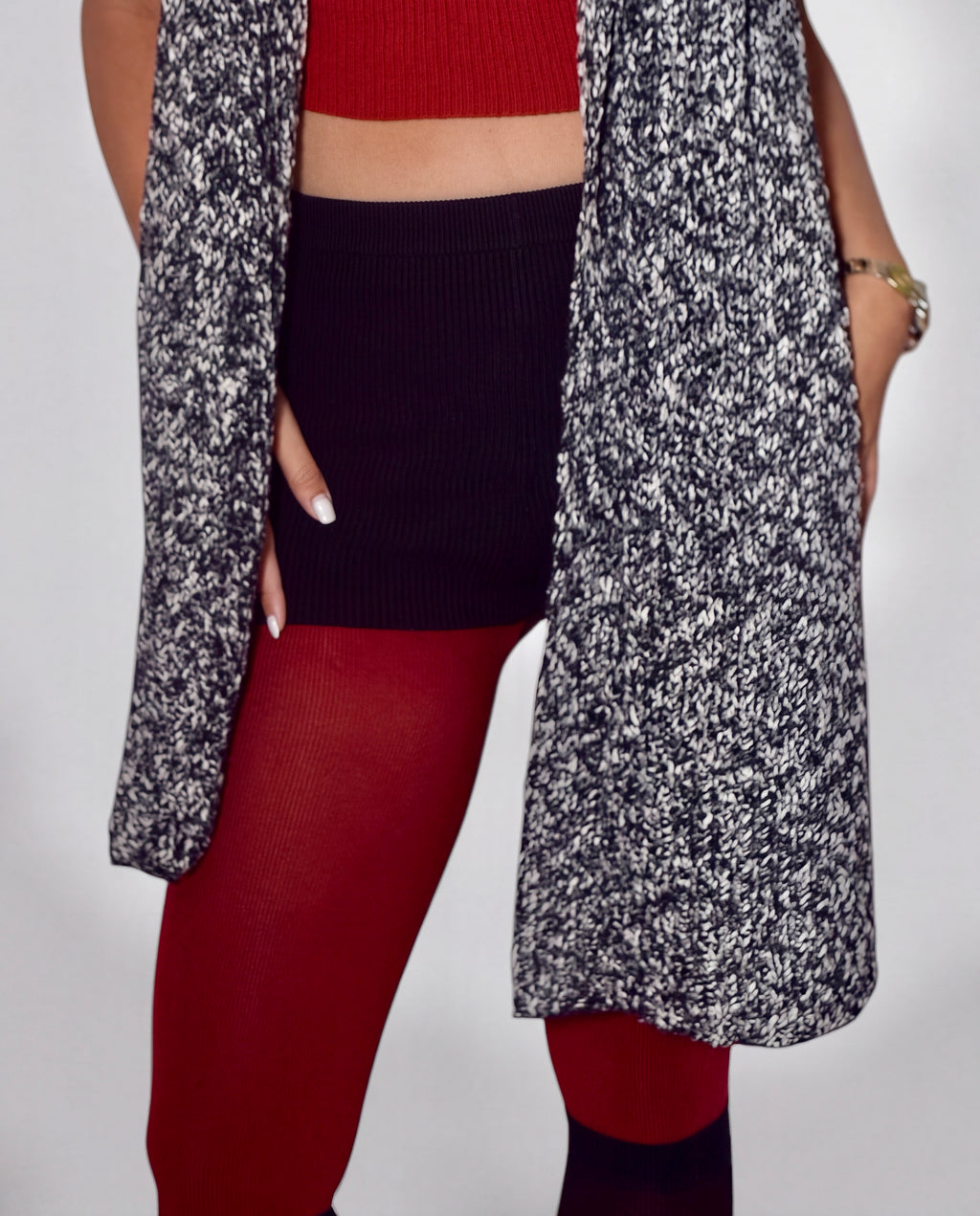 A close-up of a person wearing a long open-front cardigan with a black and white marled pattern over a red ribbed crop top and red tights, against a plain light gray studio background.