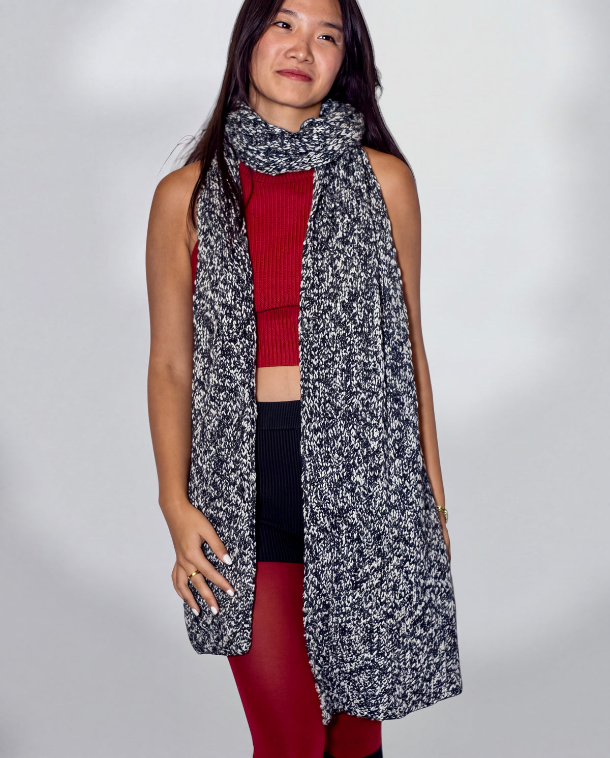 A woman modeling a long, open-front knit scarf vest with a black and white marled pattern, worn over a red ribbed crop top and black shorts.