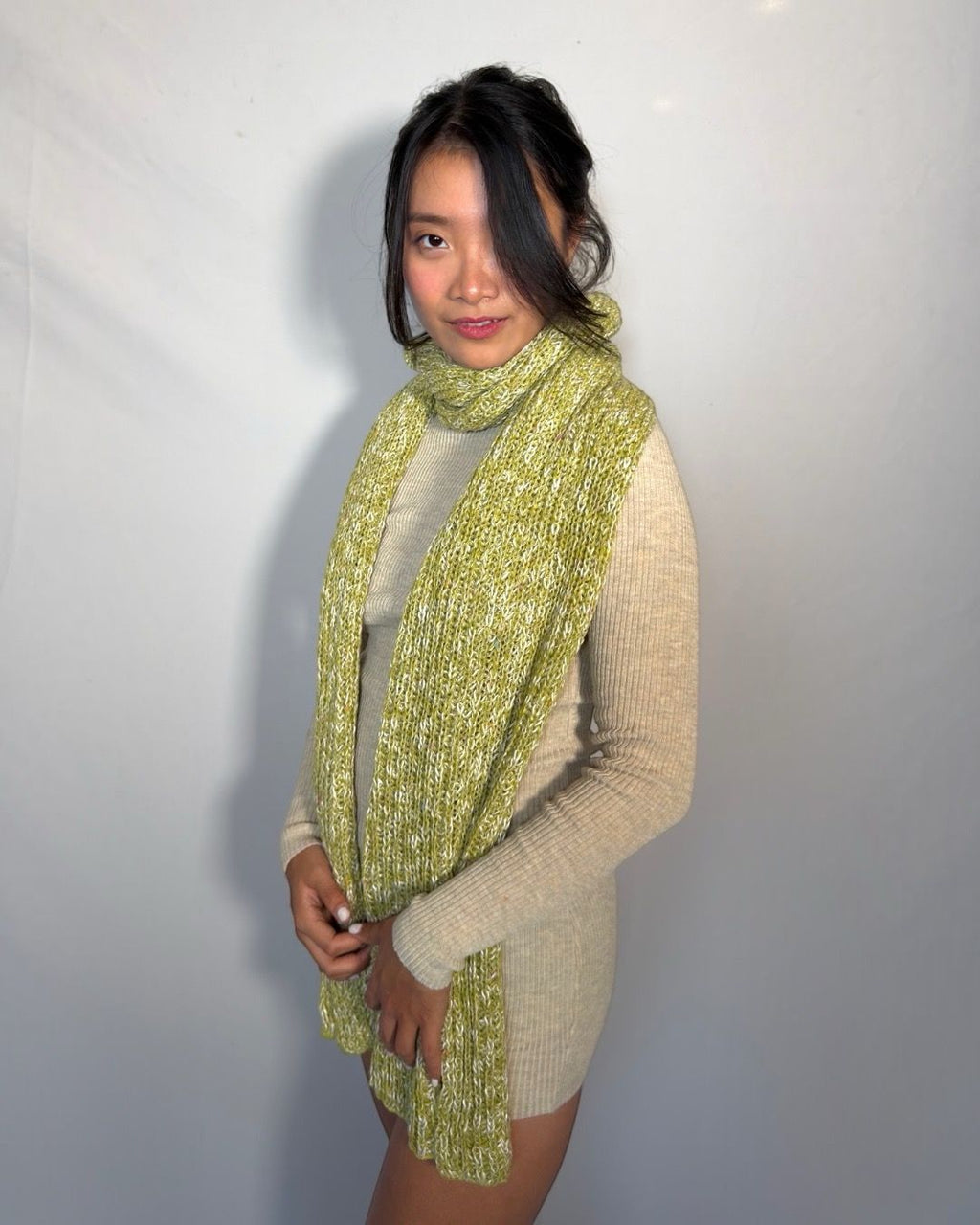 A woman modeling a chunky knit scarf in a lime green and yellow heathered pattern, worn over a beige knit sweater dress against a plain light gray background.