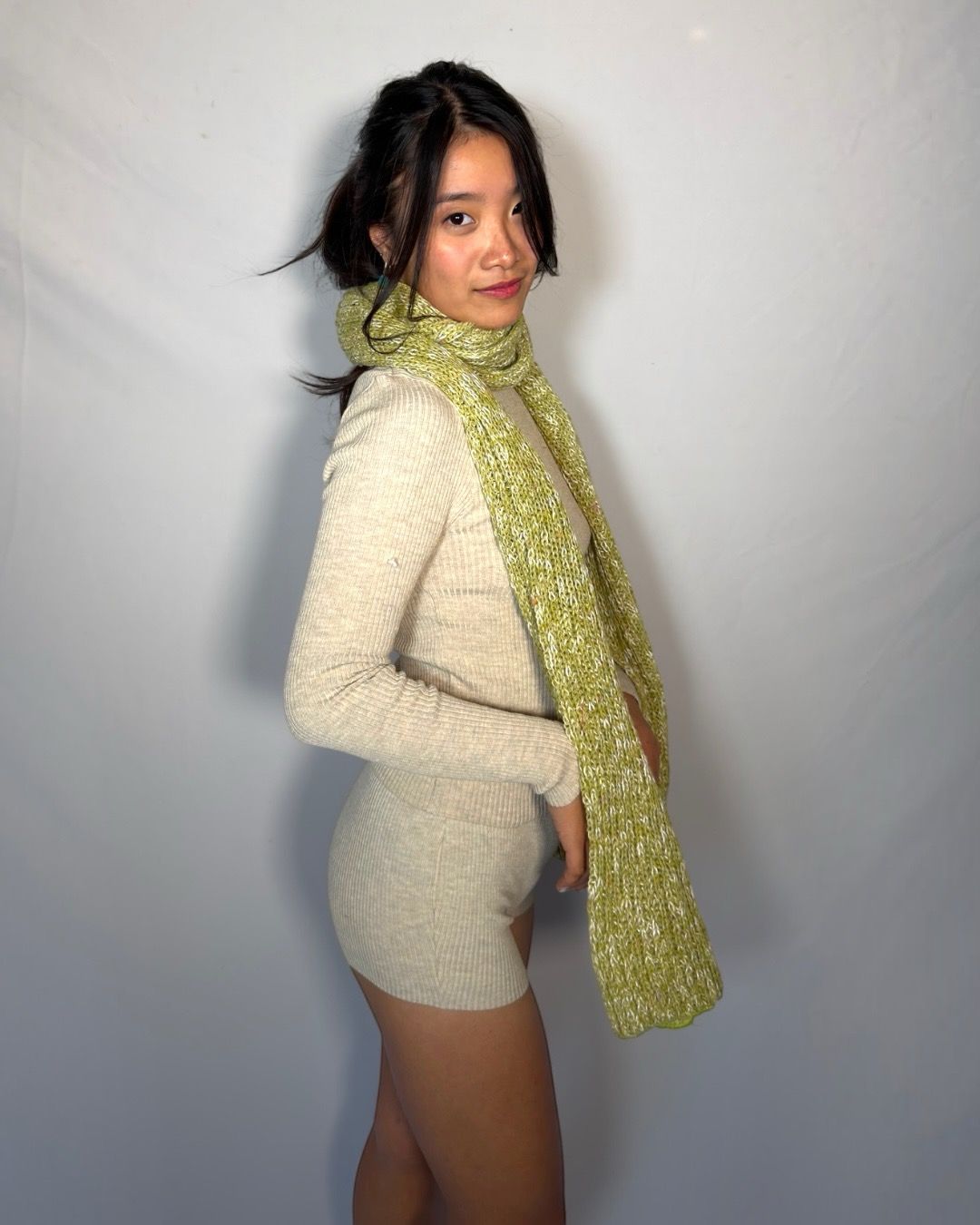 A woman wearing a cream ribbed knit mini dress and a textured lime green scarf, posing against a light gray studio backdrop.