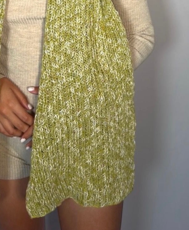 A close-up of a person wearing a chunky knit green and white marled sweater vest over a beige ribbed knit top and shorts, with the hand holding the hem of the vest.