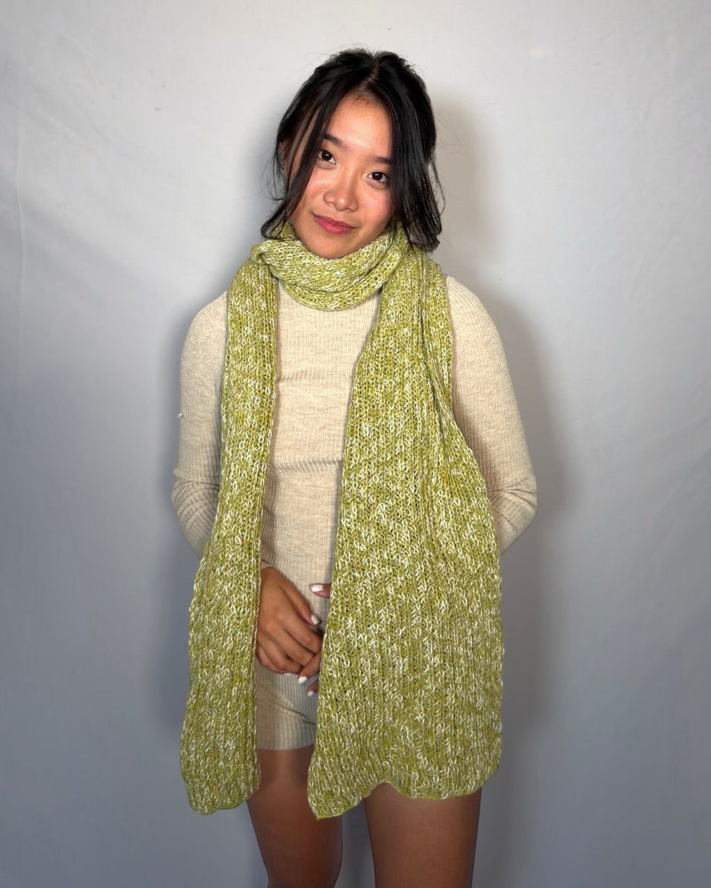 A woman wearing a chunky knit lime green scarf over a cream ribbed sweater and shorts, standing against a light gray backdrop.
