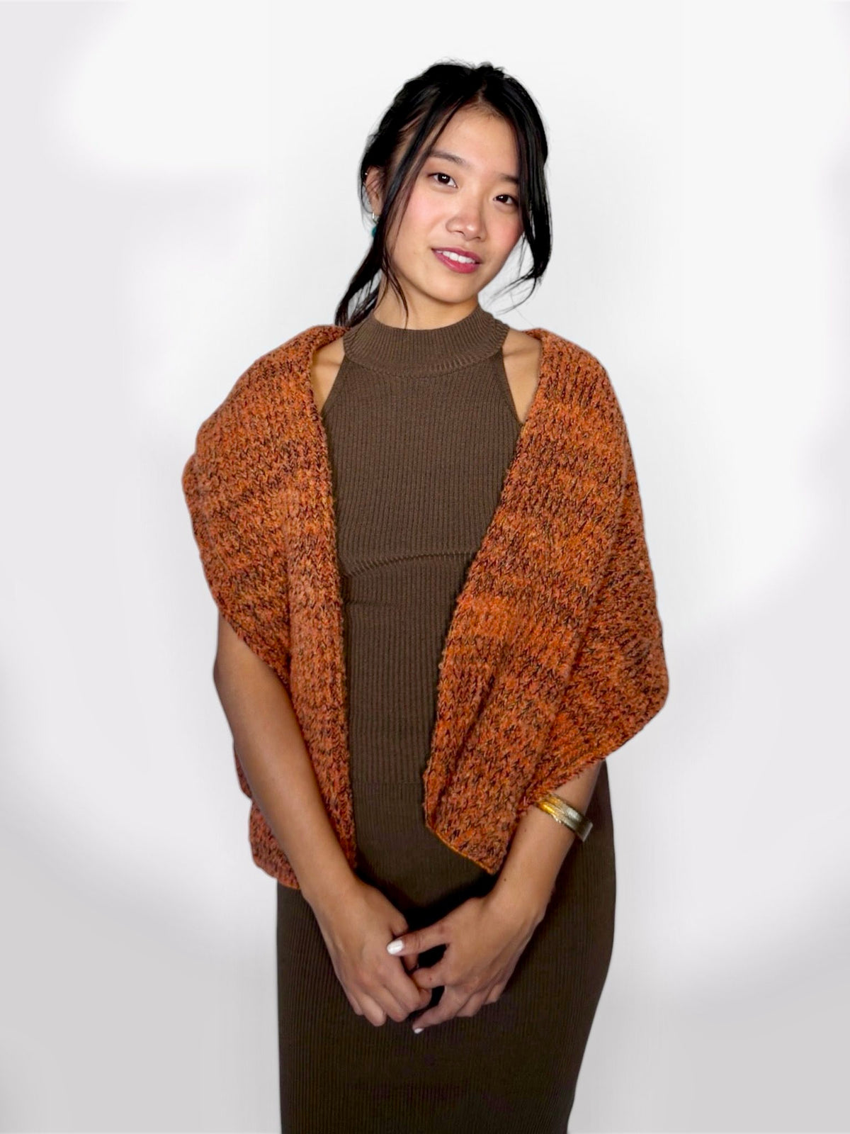 A woman modeling a rust-orange marled knit shrug or poncho over a brown ribbed mock-neck dress against a plain white studio background.
