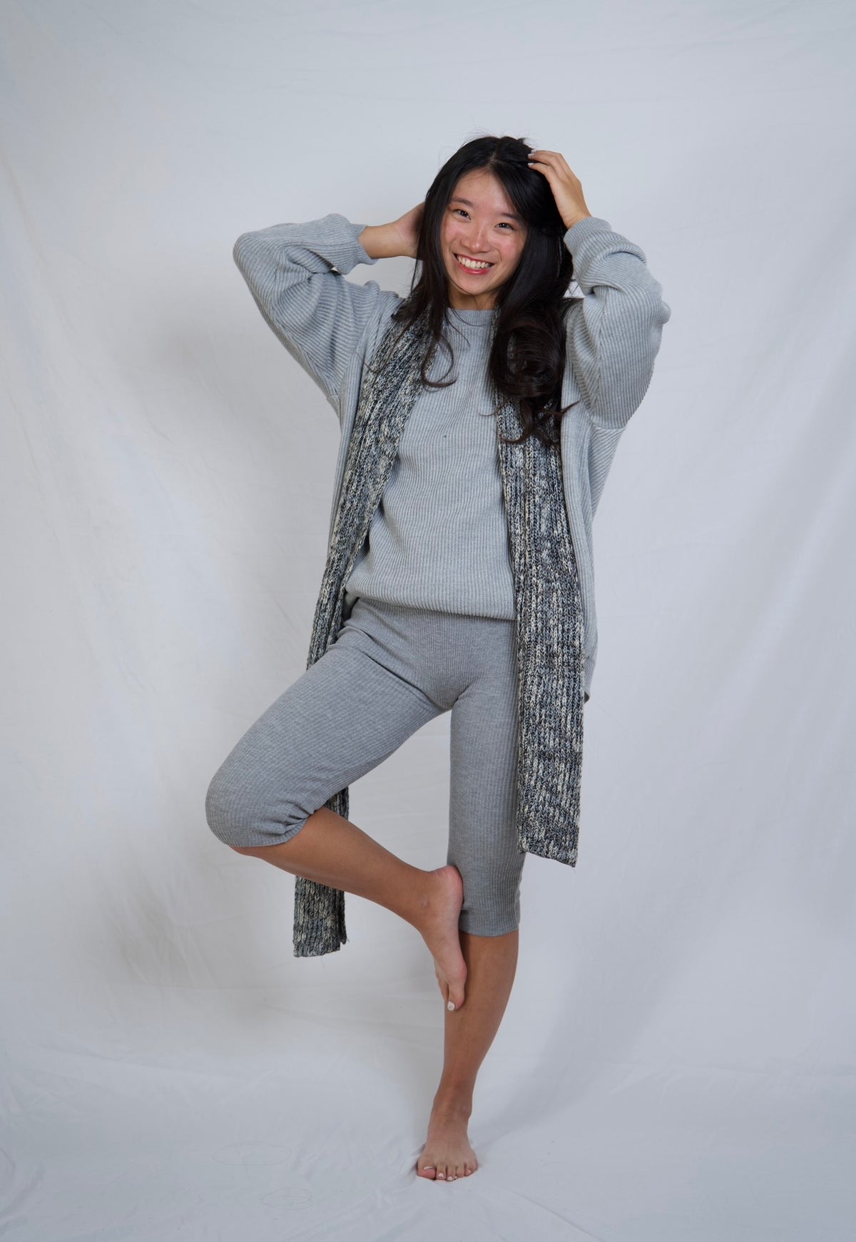 A woman posing in a gray lounge set consisting of a crewneck sweatshirt, matching capri pants, and a textured gray cardigan, set against a plain white studio backdrop.