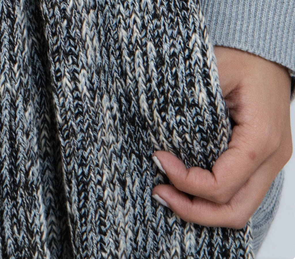 Close-up of a textured knit sweater or scarf in a heathered gray, black, and off-white pattern, with a hand holding the fabric to show its thickness and texture.
