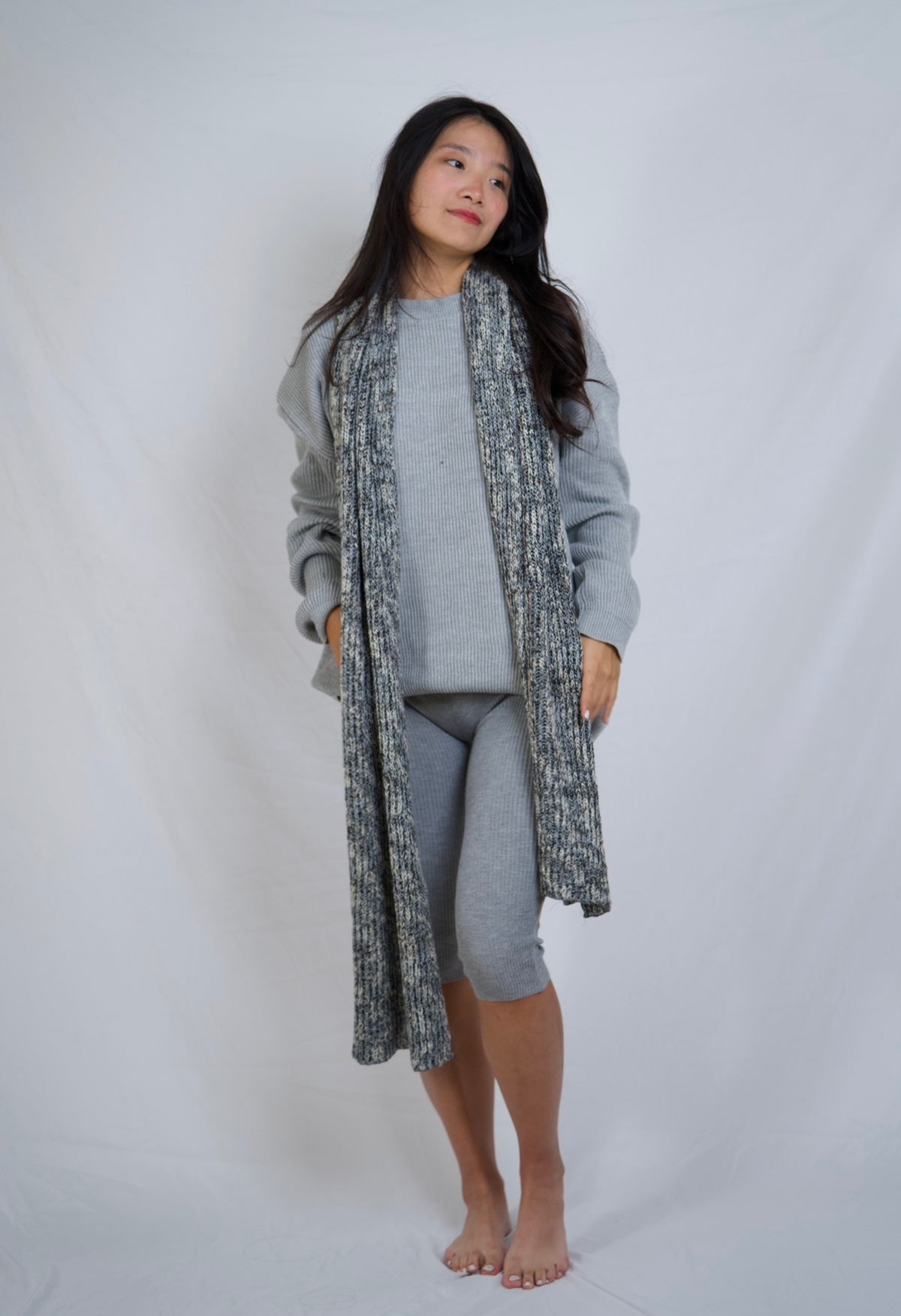 A woman modeling a gray knit lounge set consisting of a long open-front cardigan, a matching long-sleeve top, and fitted capri leggings, standing against a white studio backdrop.
