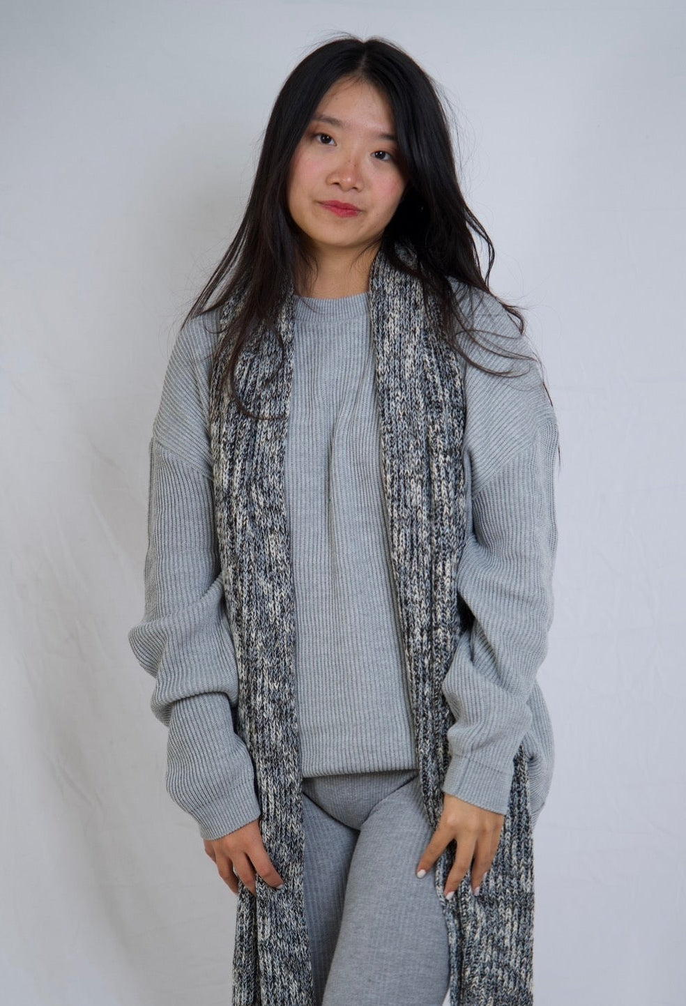A woman modeling a light gray knit matching set consisting of a crewneck sweater, fitted capri pants, and a long open-front cardigan with a textured black and gray marled pattern.