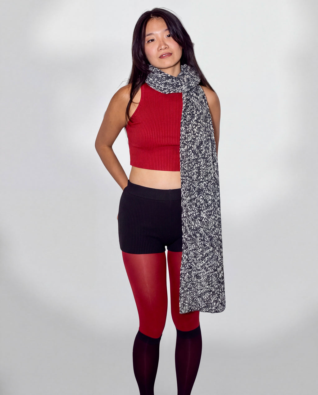 A woman modeling a black and white speckled knit scarf draped over her shoulders, paired with a red ribbed crop top, black shorts, and red tights, against a light gray studio background.