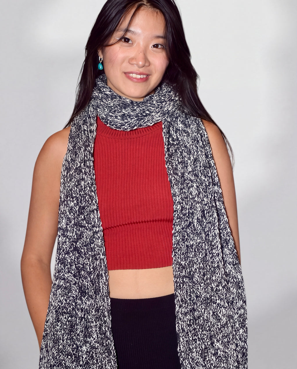 A woman wearing a black and white marled knit scarf over a red ribbed crop top and black pants, photographed against a plain light gray background.