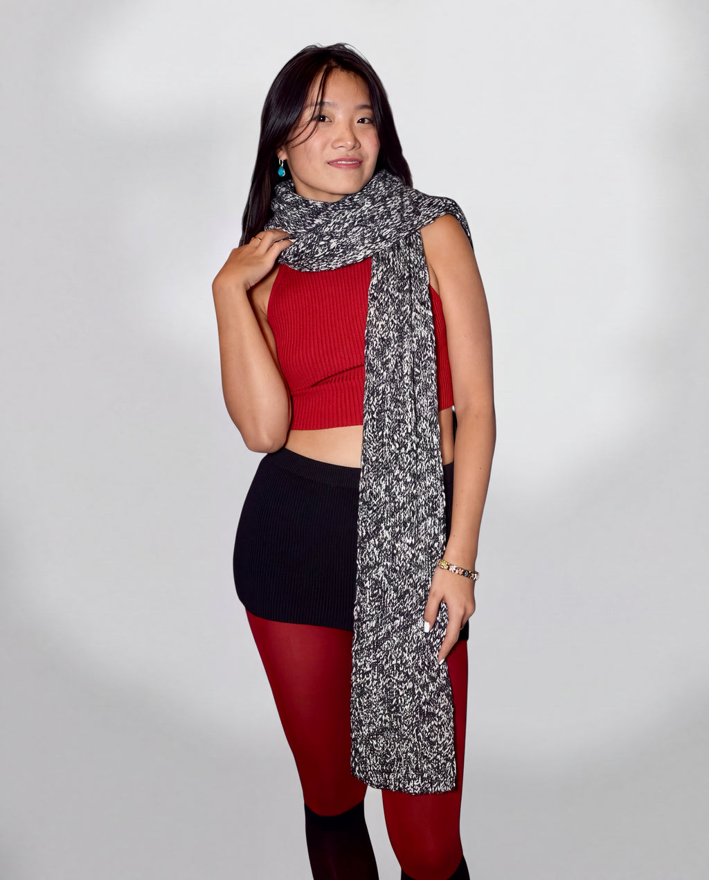 A woman modeling a long, black and white marled knit scarf draped over her shoulders, paired with a red ribbed crop top, black skirt, and red tights, against a light gray studio background.