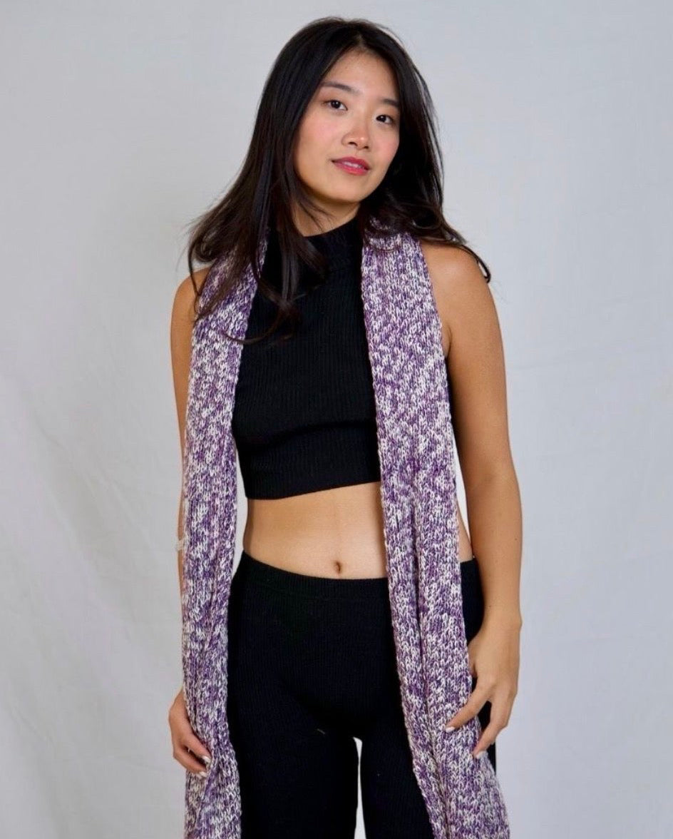 A woman modeling a long, sleeveless open-front cardigan with a purple and white abstract pattern, worn over a black crop top and black pants against a light gray studio backdrop.