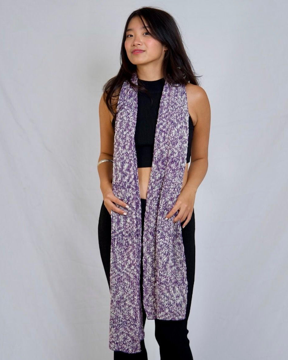 A woman modeling a long, open-front scarf with a purple and white abstract pattern, styled over a black crop top and black pants against a light gray studio backdrop.