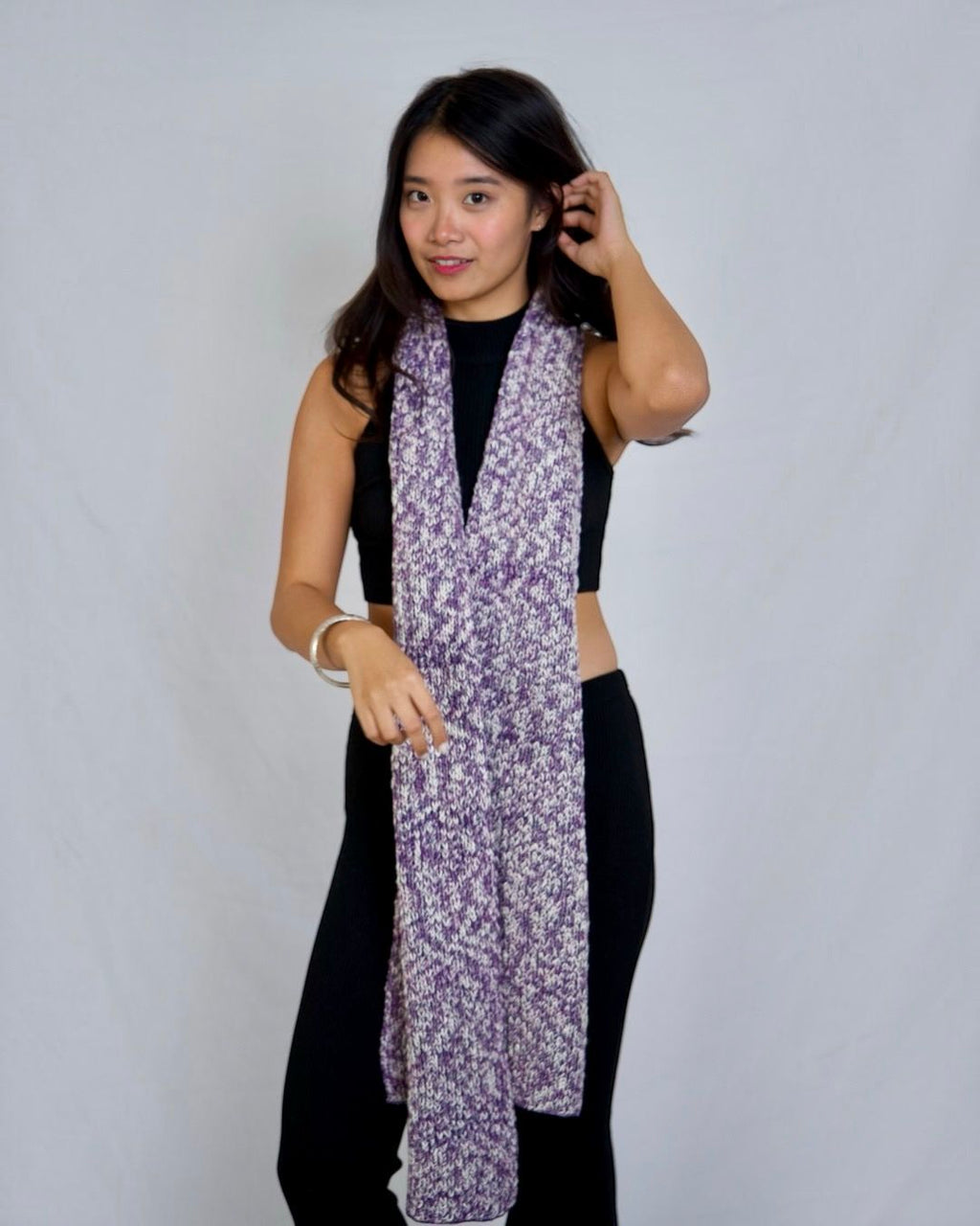 A woman modeling a lightweight purple and white patterned scarf draped over her shoulders, paired with a black crop top and black pants, against a light gray studio background.