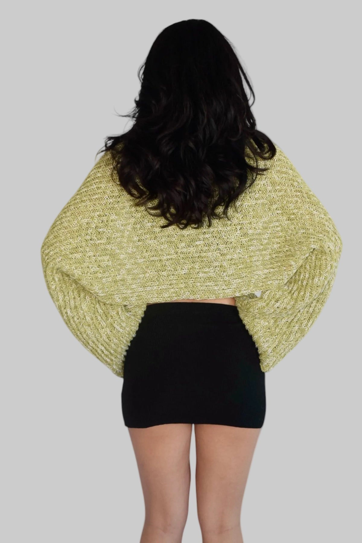 A woman viewed from behind wearing a chunky, textured knit shrug or cape in a light olive green color, paired with a black mini skirt, against a plain white background.