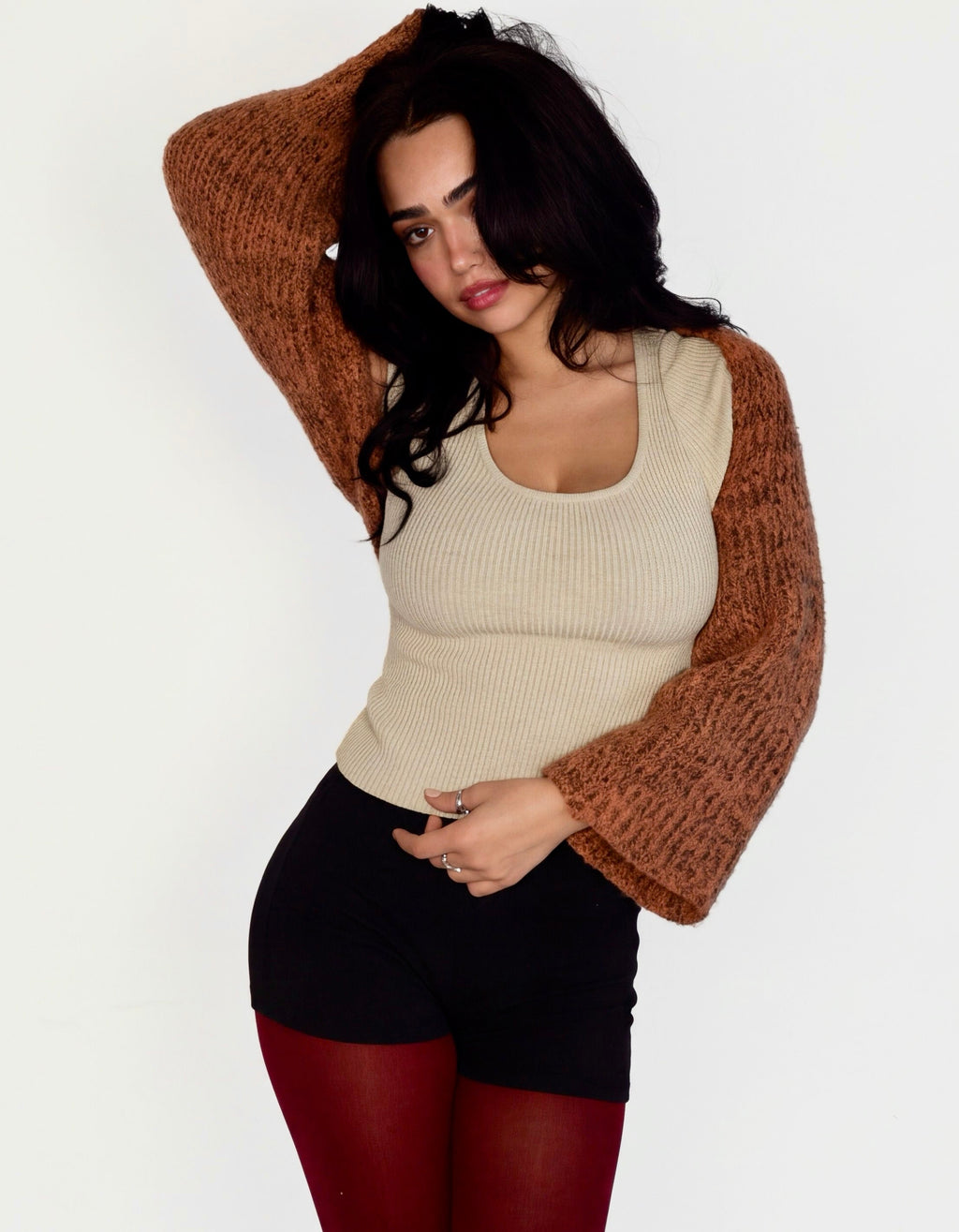 A woman modeling a beige ribbed tank top with brown knit sleeves, black shorts, burgundy tights, and black platform shoes against a plain white studio background.