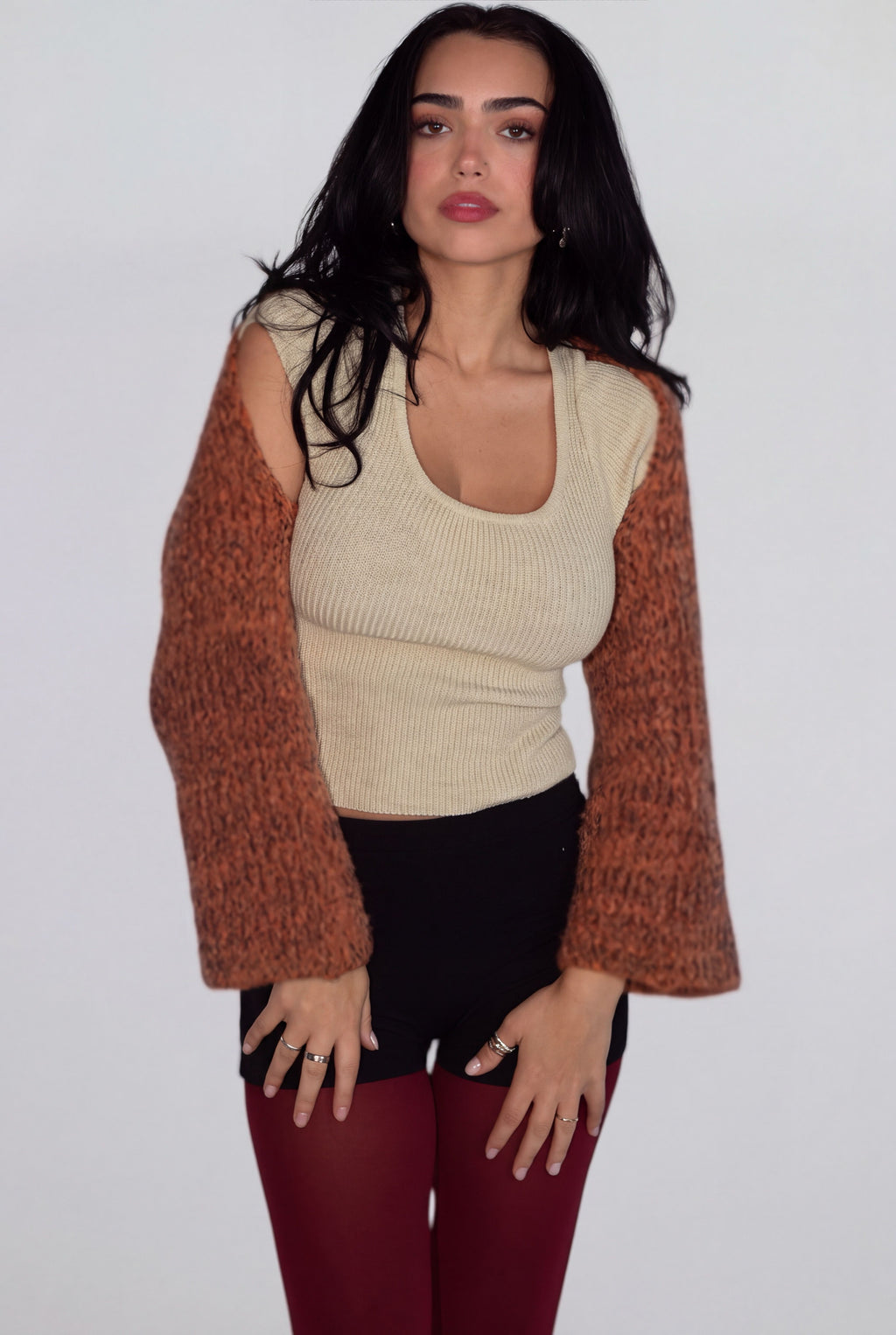 A woman modeling a cream ribbed knit crop top with long, burnt orange shrug sleeves, paired with black shorts and burgundy leggings, against a plain light gray background.