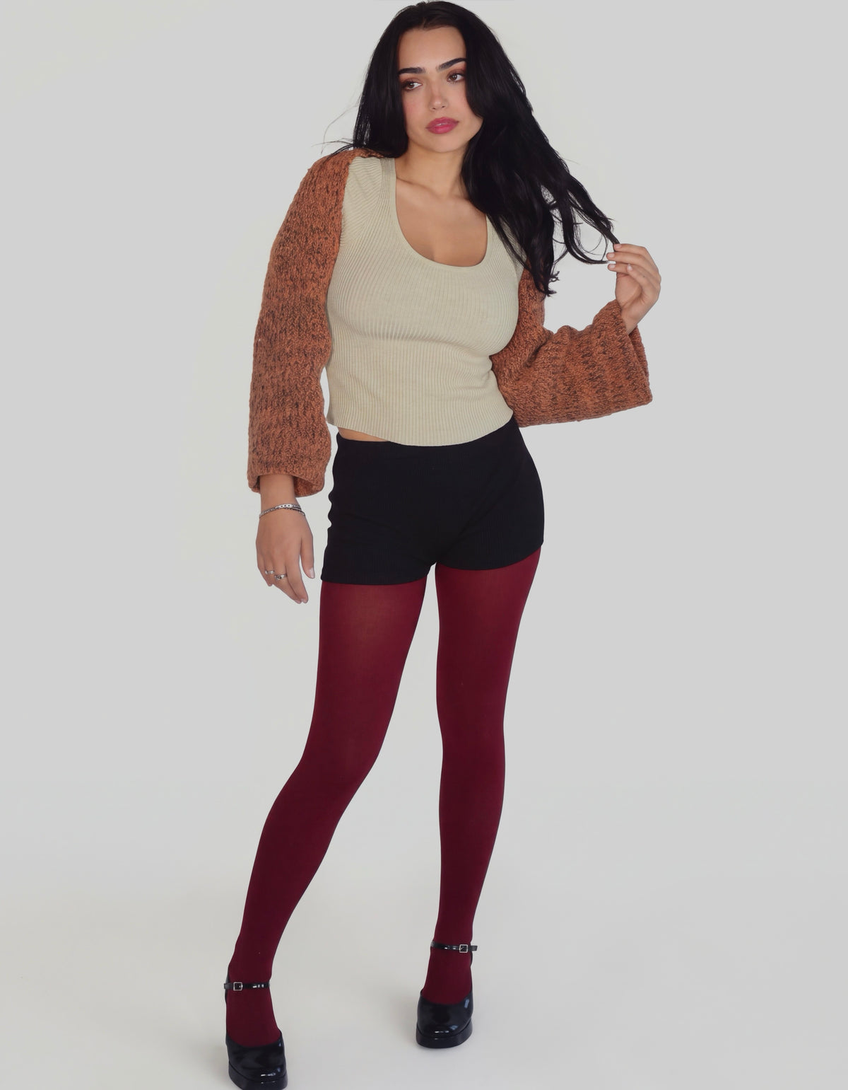 A woman modeling a knit cardigan with a beige body and rust-brown sleeves, paired with a cream tank top, black shorts, burgundy tights, and black Mary Jane heels against a light gray studio background.