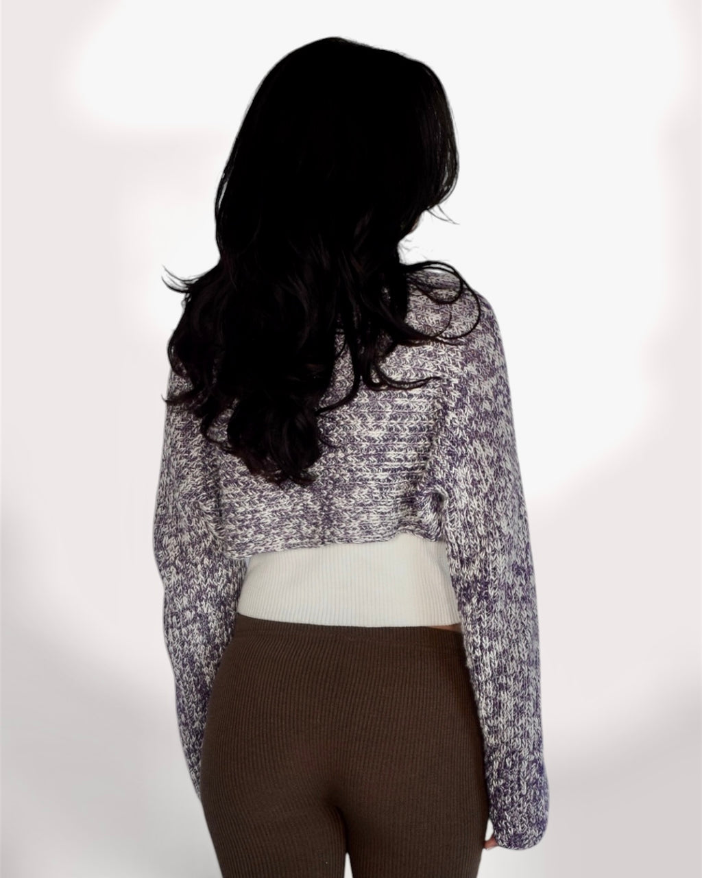 A woman shown from the back wearing a long-sleeve cropped knit sweater with a mottled purple and white pattern, layered over a cream ribbed top and brown ribbed leggings, against a plain white background.