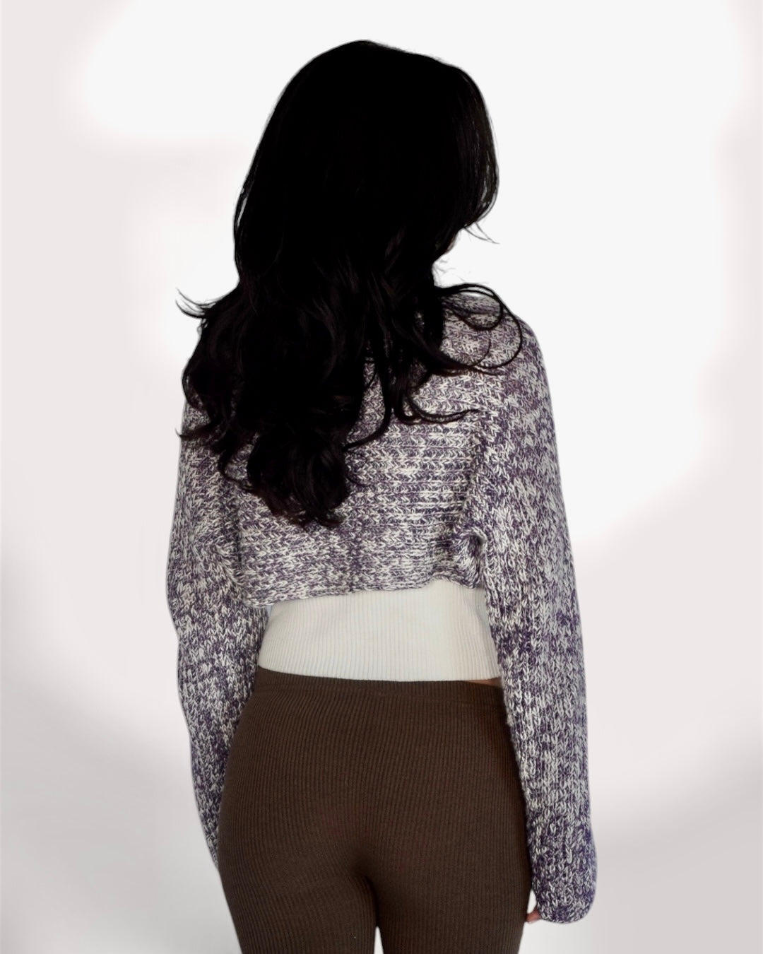 A woman shown from the back wearing a long-sleeve cropped knit sweater with a mottled purple and white pattern, layered over a cream ribbed top and brown ribbed leggings, against a plain white background.