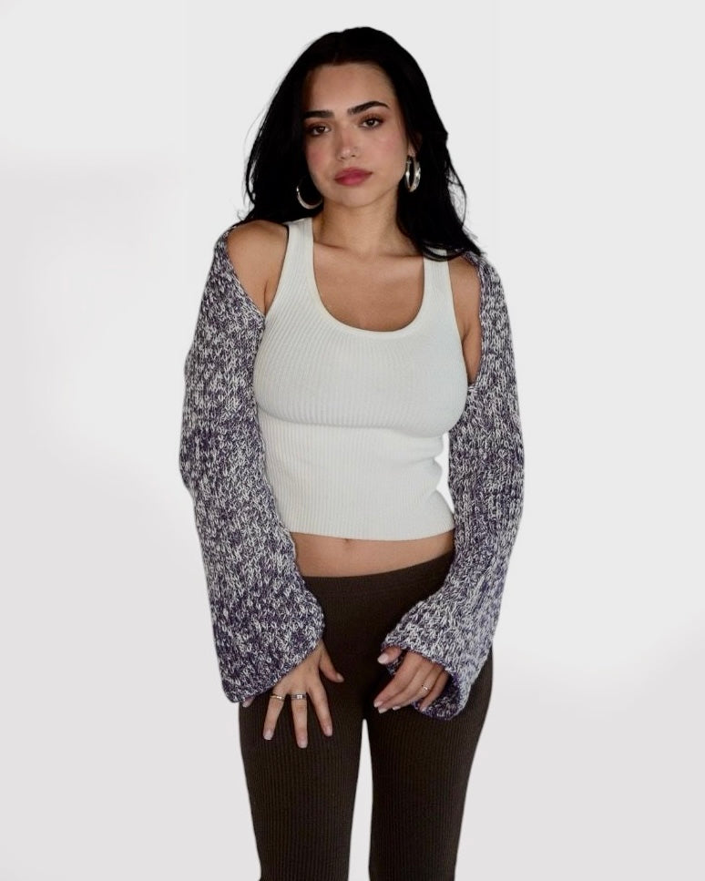 A woman modeling a white ribbed tank top with long sleeves, worn open over a marled gray and purple knit cardigan, paired with dark brown ribbed pants, against a plain light gray background.