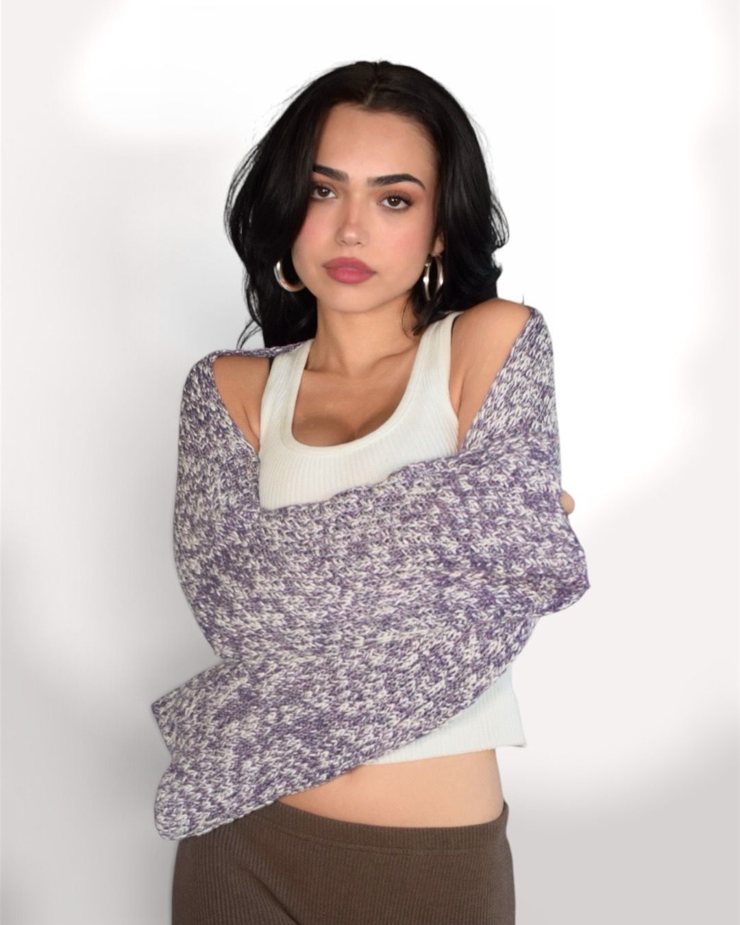 A woman modeling a lavender and white marled knit shawl draped over her shoulders, worn over a white ribbed tank top and brown pants, against a plain white background.