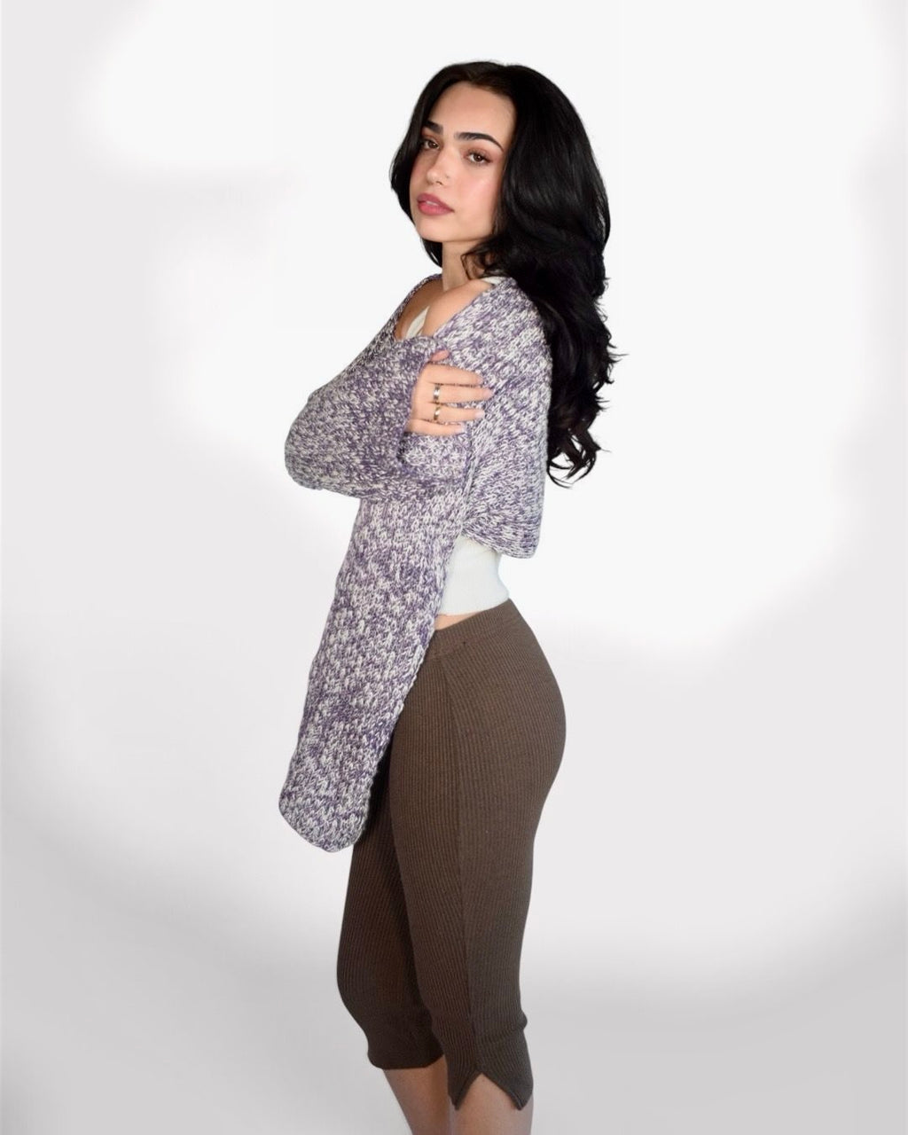 A woman modeling a long-sleeve, open-front knit cardigan with a heathered purple and white pattern, worn over a white ribbed tank top and brown ribbed knee-length pants, set against a plain light gray studio background.