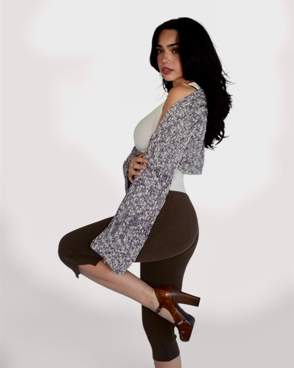 A woman posing in a studio setting, wearing a gray and white patterned cardigan over a white top, brown ribbed capri leggings, and brown heeled mules.