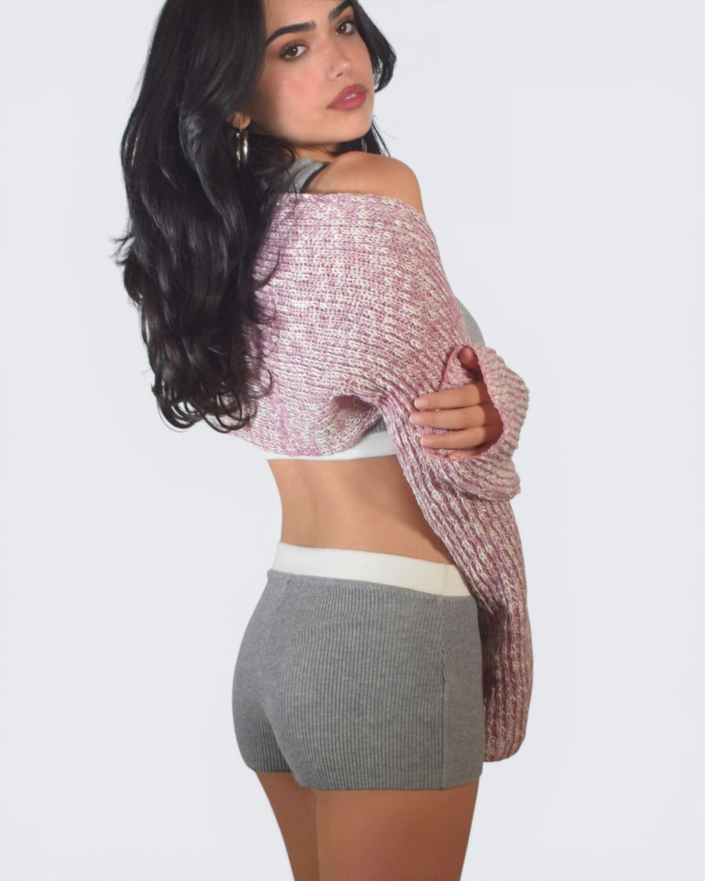 Woman wearing a pink knit cardigan over a white top and gray shorts on a plain background