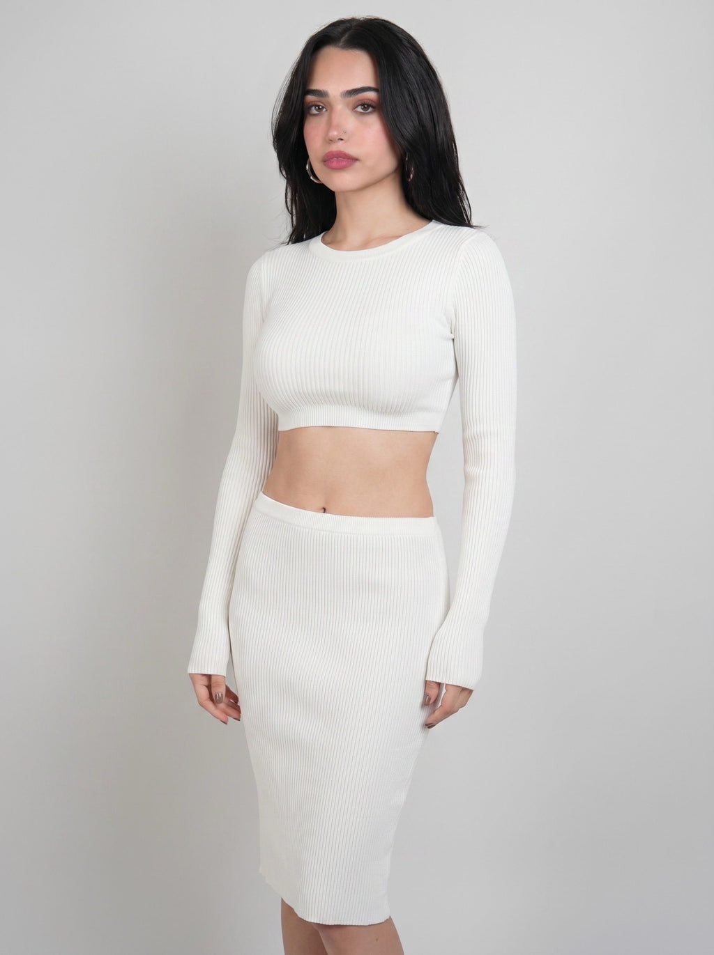 A woman modeling a white ribbed knit two-piece set consisting of a long-sleeve crop top and a matching high-waisted pencil skirt, set against a plain light gray studio background.