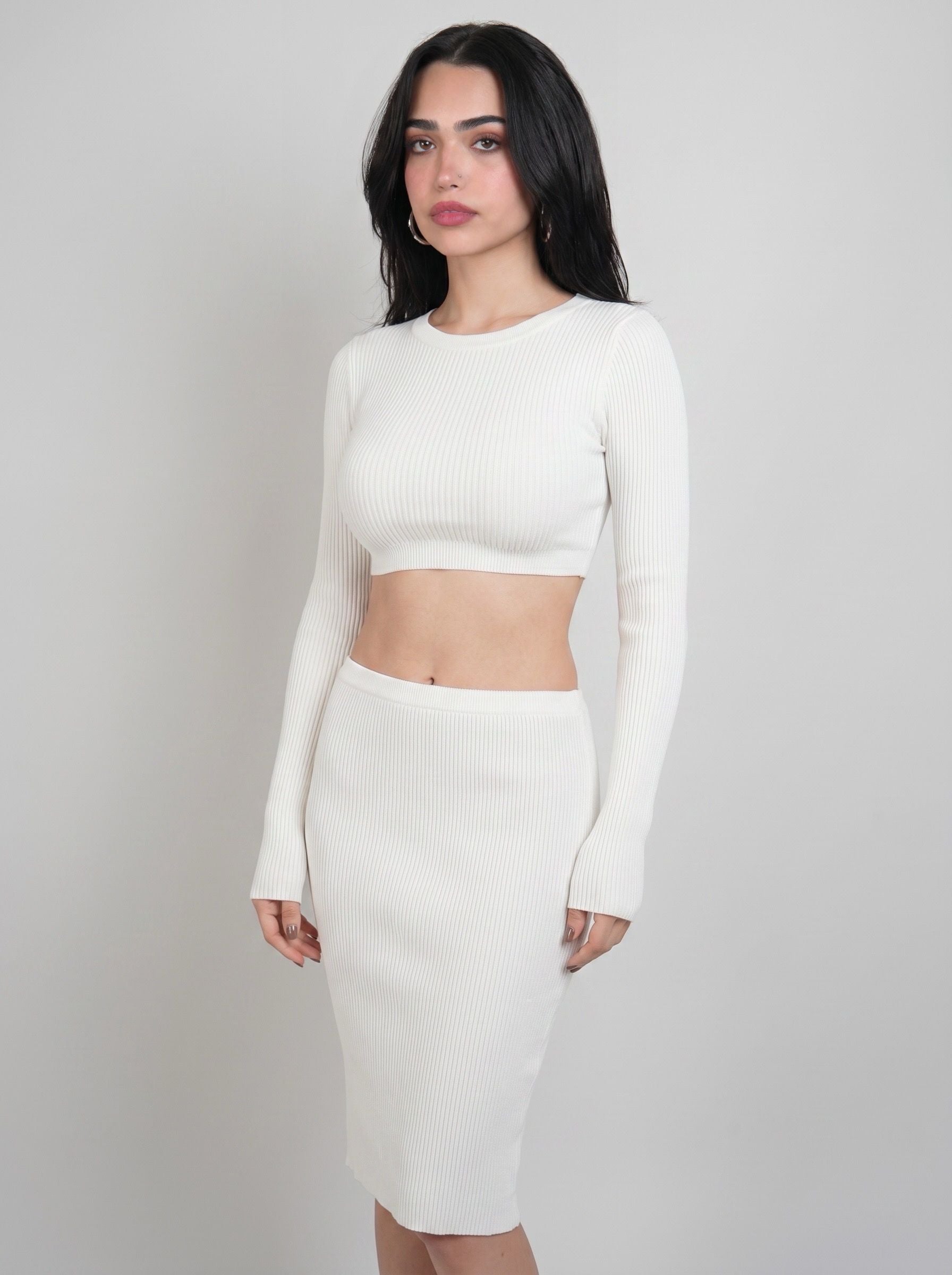 A woman modeling a white ribbed knit two-piece set consisting of a long-sleeve crop top and a matching high-waisted pencil skirt, set against a plain light gray studio background.