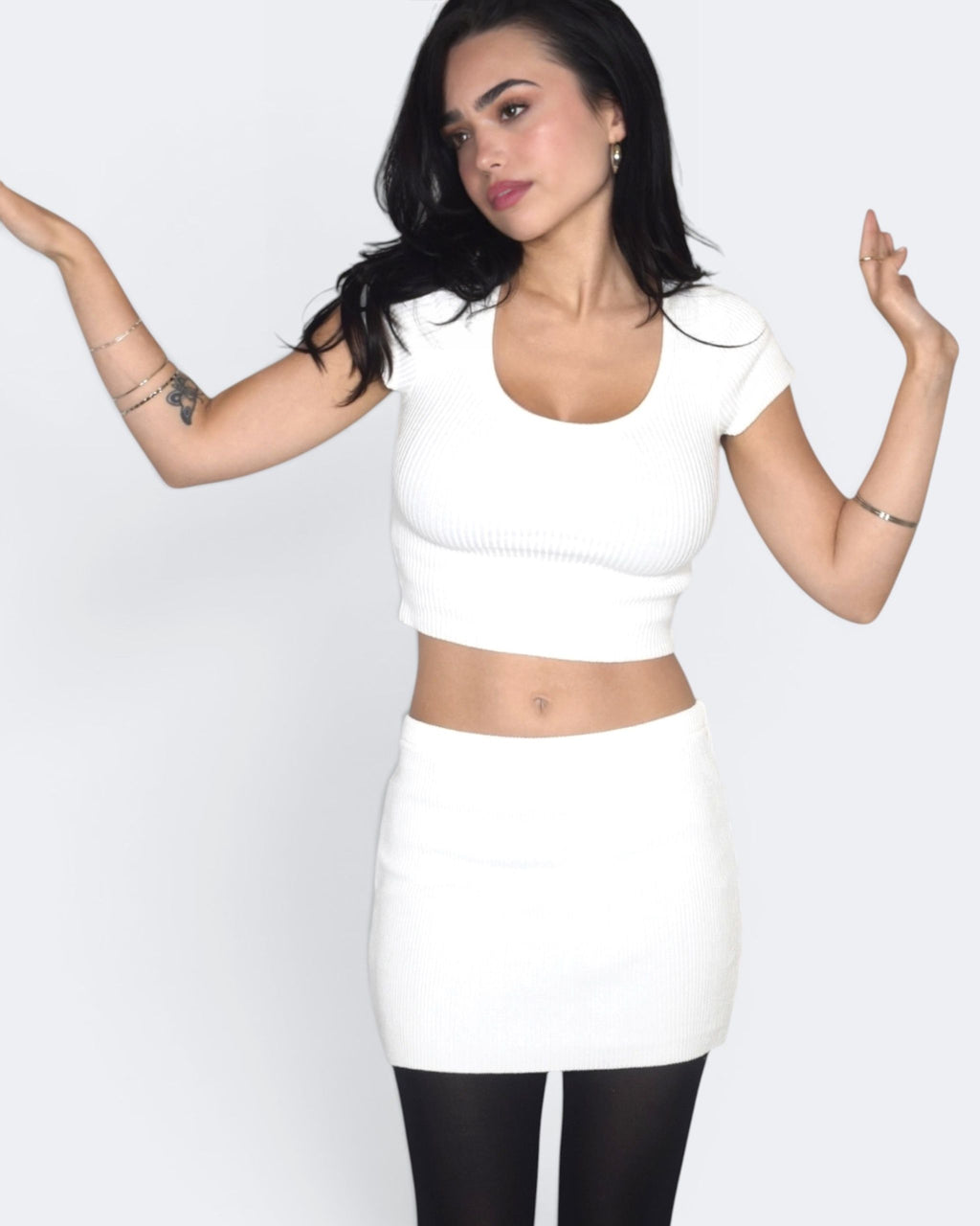 Woman wearing a white crop top and skirt with black tights on a plain background